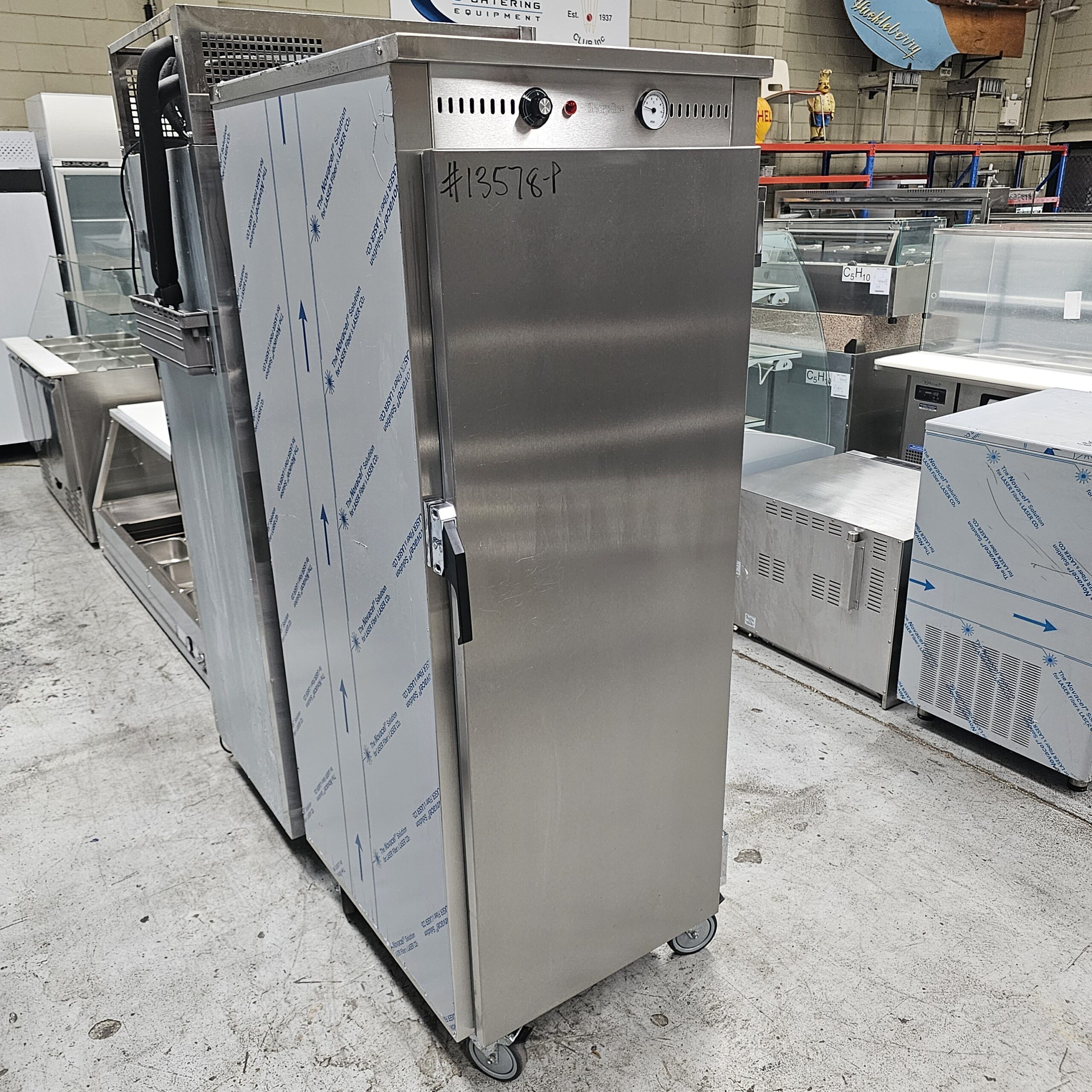 A tall, stainless steel commercial kitchen appliance on wheels stands in a warehouse with other metal equipment. The appliance has a control panel with dials and a handwritten number #125789 on its door. MRCE
