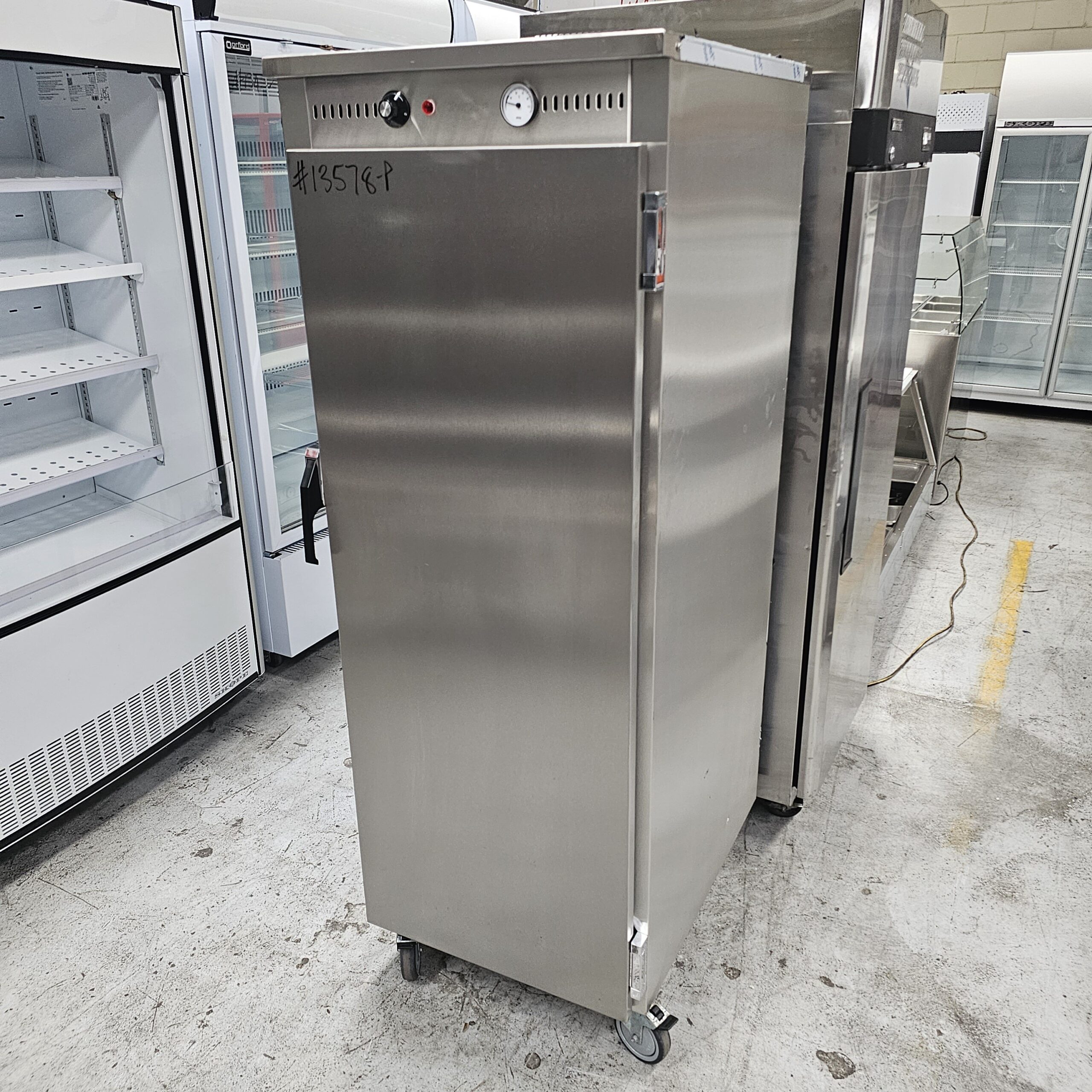 A stainless steel commercial food warming cabinet on wheels stands in a warehouse with refrigerators and empty display cases in the background. The cabinet has a thermometer and a handle on the front door. MRCE