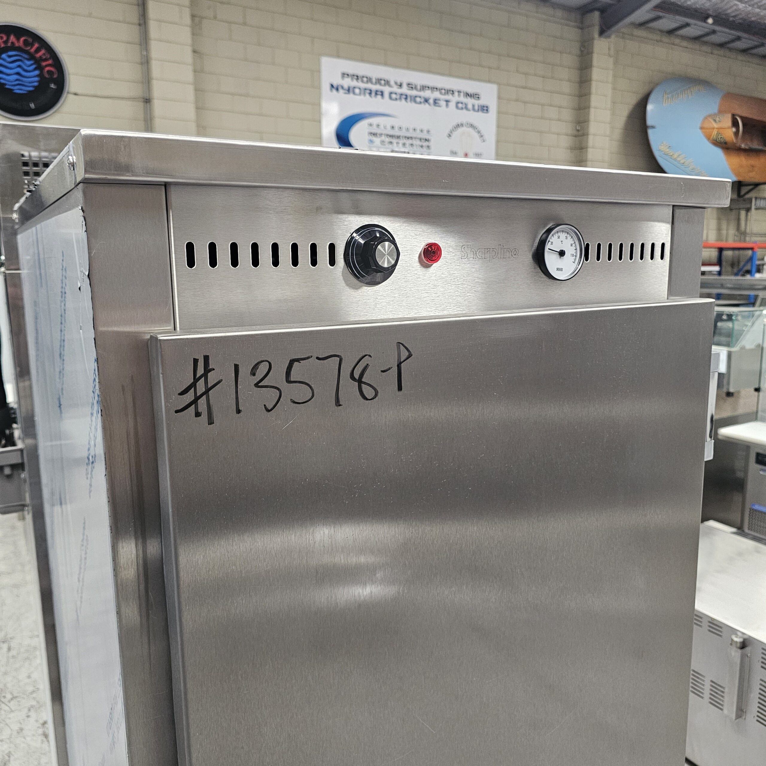 A stainless steel commercial appliance with a thermostat, gauge, and red indicator light on top. The number #13578-P is written on the front. Other equipment and a Nyora Cricket Club sign are visible in the background. MRCE