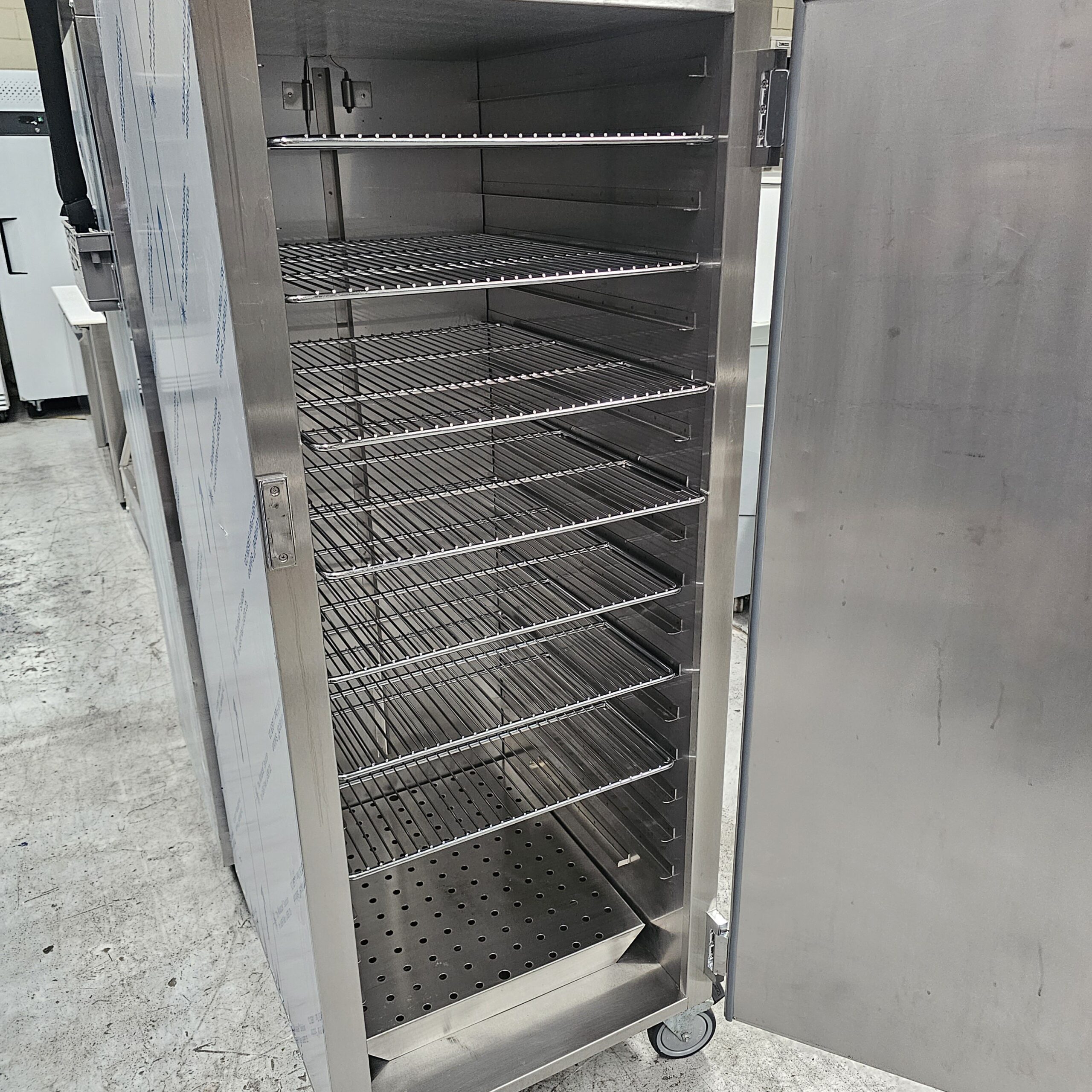 A large stainless steel commercial food storage rack with multiple wire shelves and a perforated bottom, standing on wheels with its door open in an industrial setting. MRCE