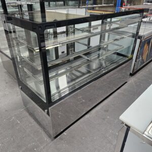 A large, empty glass display case with multiple shelves, set on a shiny metal base, stands on a concrete floor in what appears to be a warehouse or showroom. MRCE