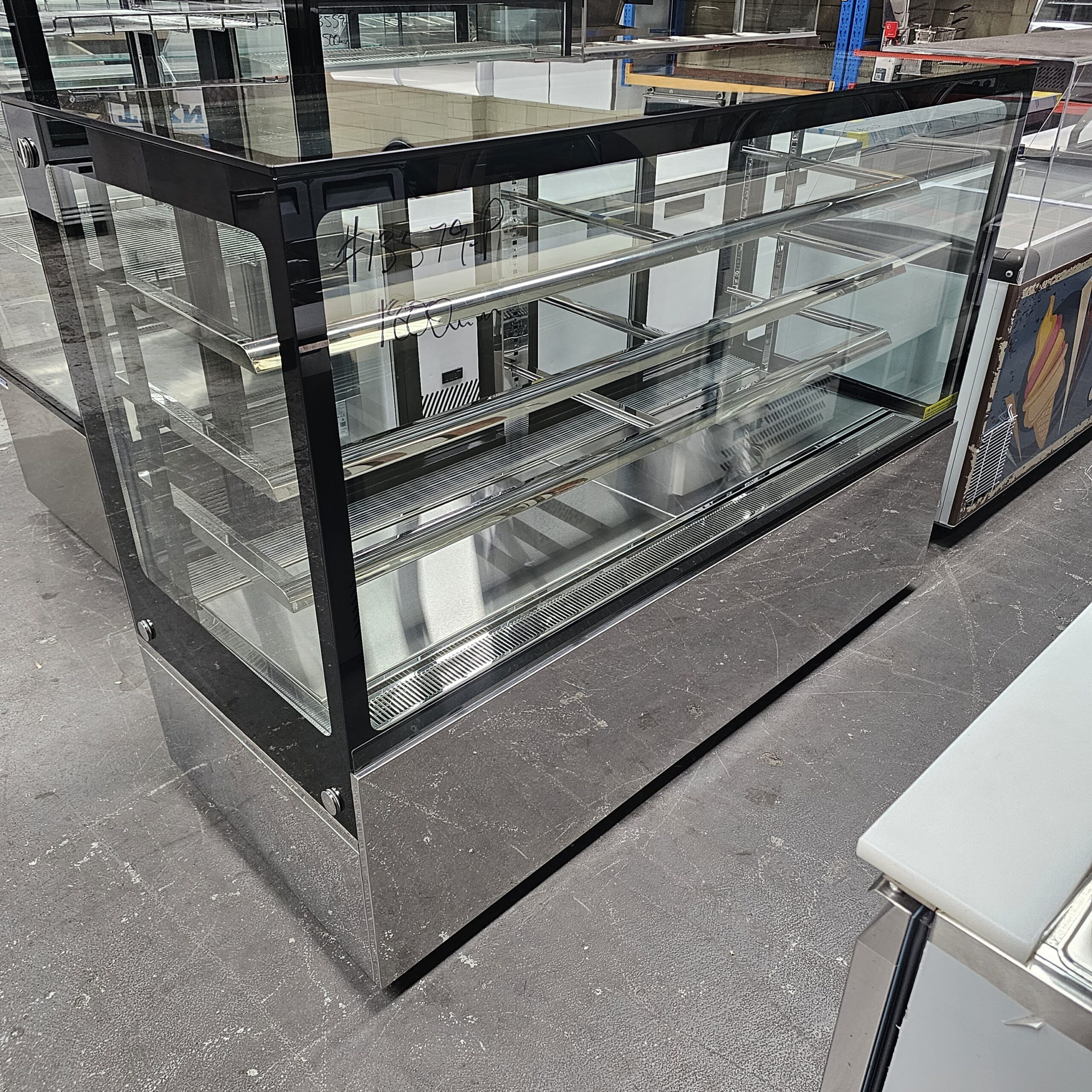 A large, empty glass display case with multiple shelves, set on a shiny metal base, stands on a concrete floor in what appears to be a warehouse or showroom. MRCE