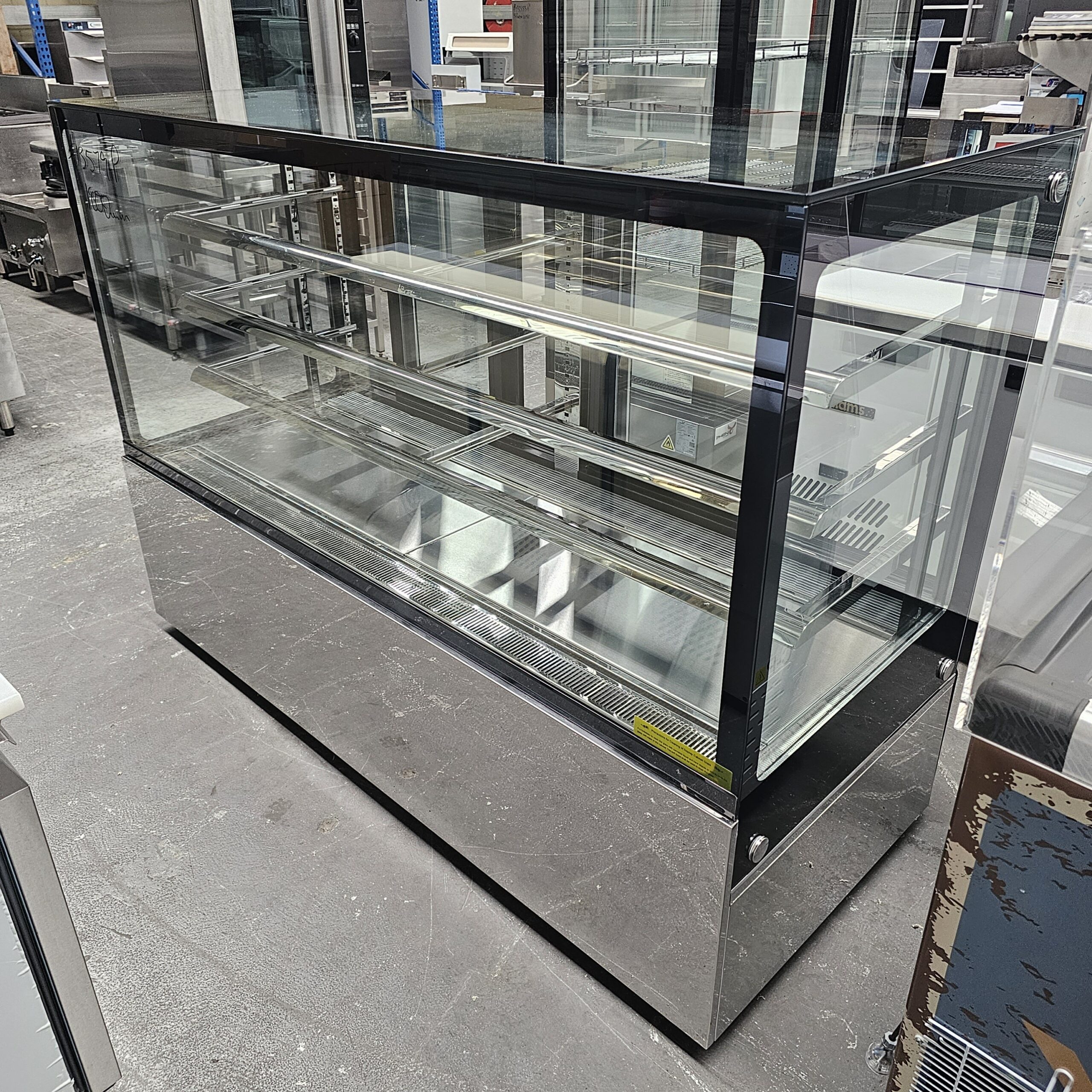 A large, empty commercial glass display case with multiple shelves and a stainless steel base sits on a concrete floor in a showroom or warehouse. MRCE
