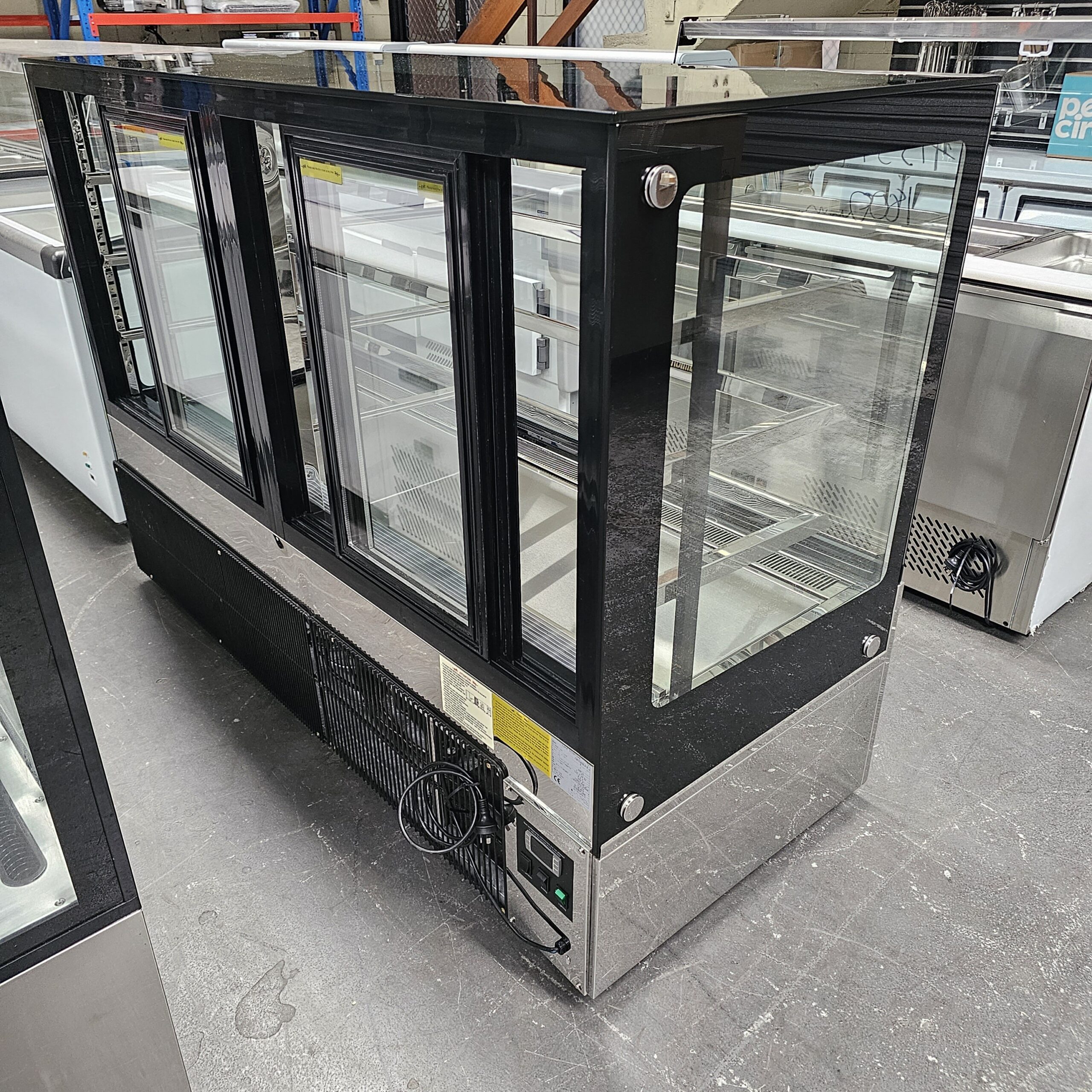 A commercial glass display refrigerator with sliding doors, black frame, and stainless steel base stands on a concrete floor in a warehouse setting. Shelves inside are empty. MRCE