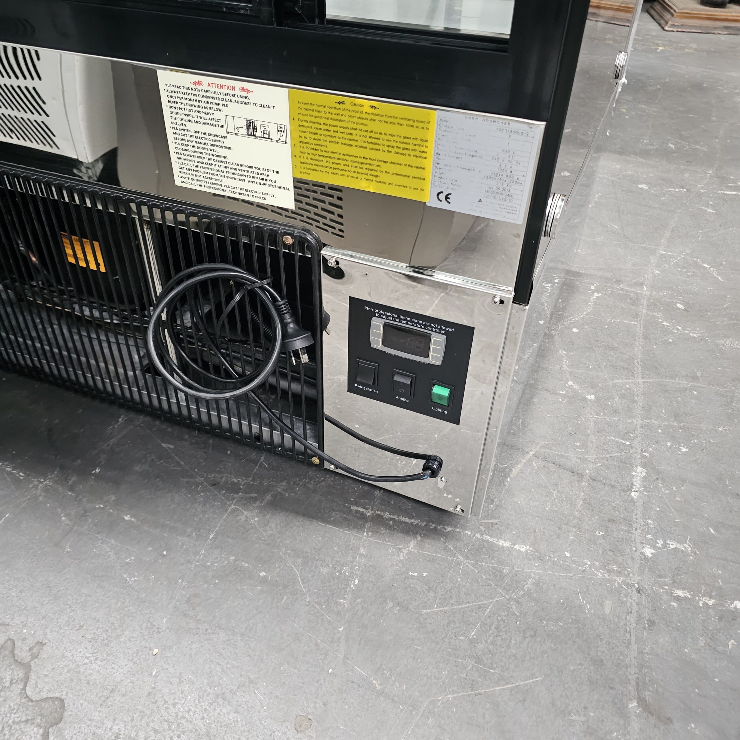 Close-up of the lower side of a commercial refrigerator, showing a control panel with switches, a display screen, a coiled power cord, ventilation grills, and instruction labels on a metallic surface. MRCE