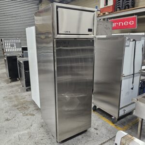 A stainless steel commercial refrigerator stands in a warehouse among other kitchen equipment, with handwritten text on the door and industrial shelves visible in the background. MRCE