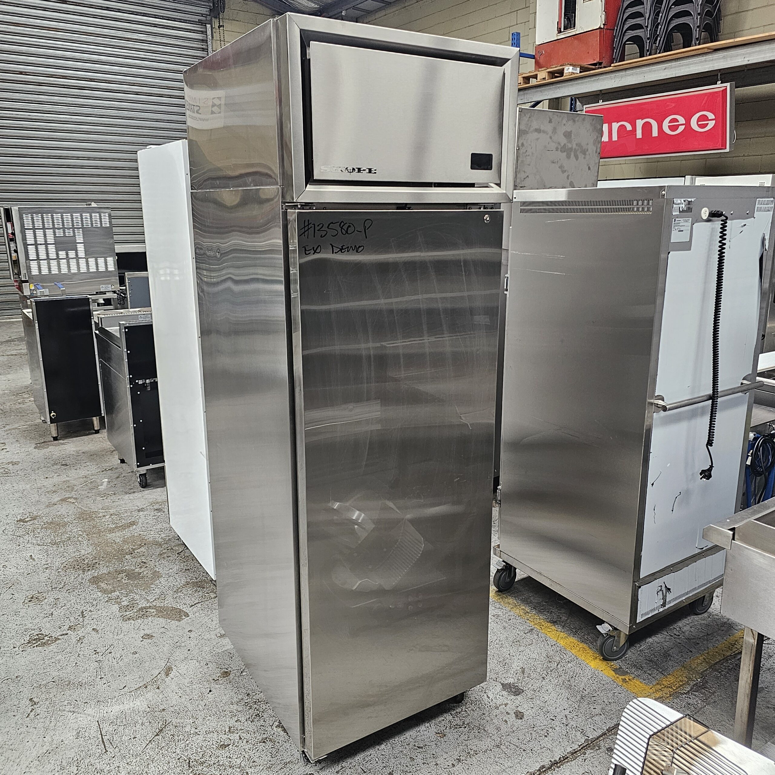 A stainless steel commercial refrigerator stands in a warehouse among other kitchen equipment, with handwritten text on the door and industrial shelves visible in the background. MRCE