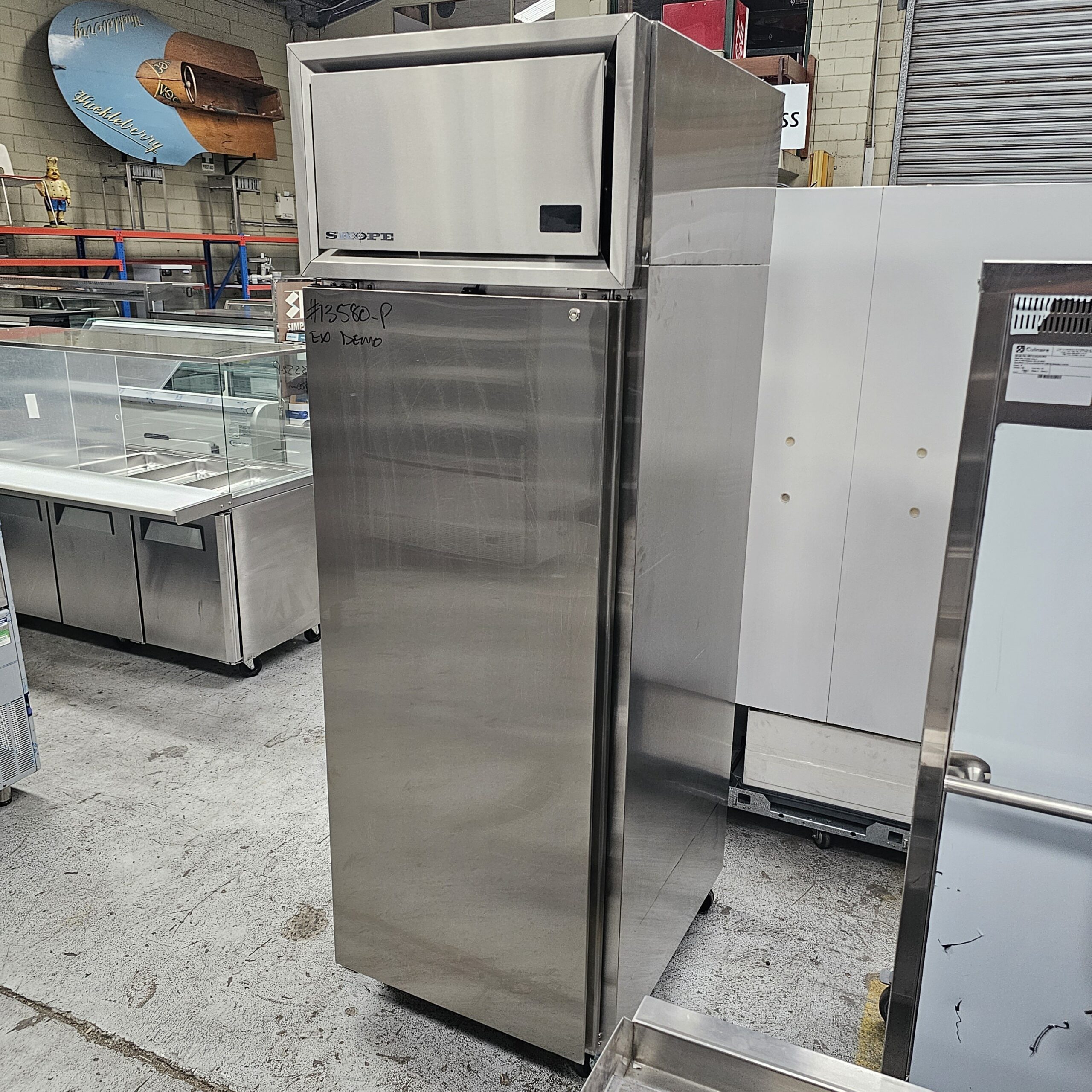 A tall, stainless steel commercial refrigerator stands in a warehouse with various equipment and shelves visible in the background. Handwritten text is visible on the front door. MRCE