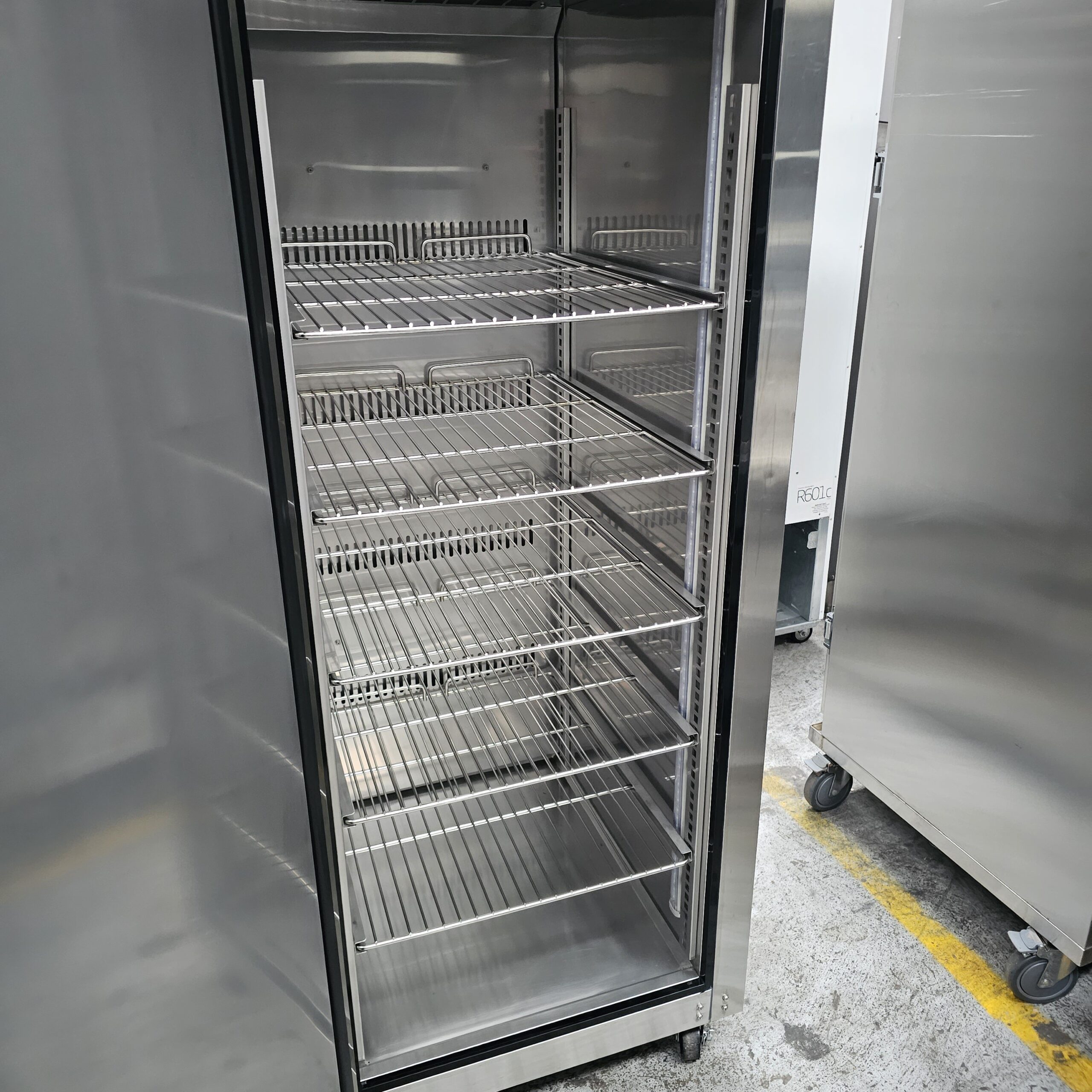 A large, industrial stainless steel refrigerator with its door open, revealing empty metal shelves inside. The unit is situated on a concrete floor with another similar refrigerator nearby. MRCE