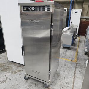 A stainless steel commercial food warmer or holding cabinet on wheels is positioned on a warehouse floor, surrounded by other industrial kitchen equipment. MRCE