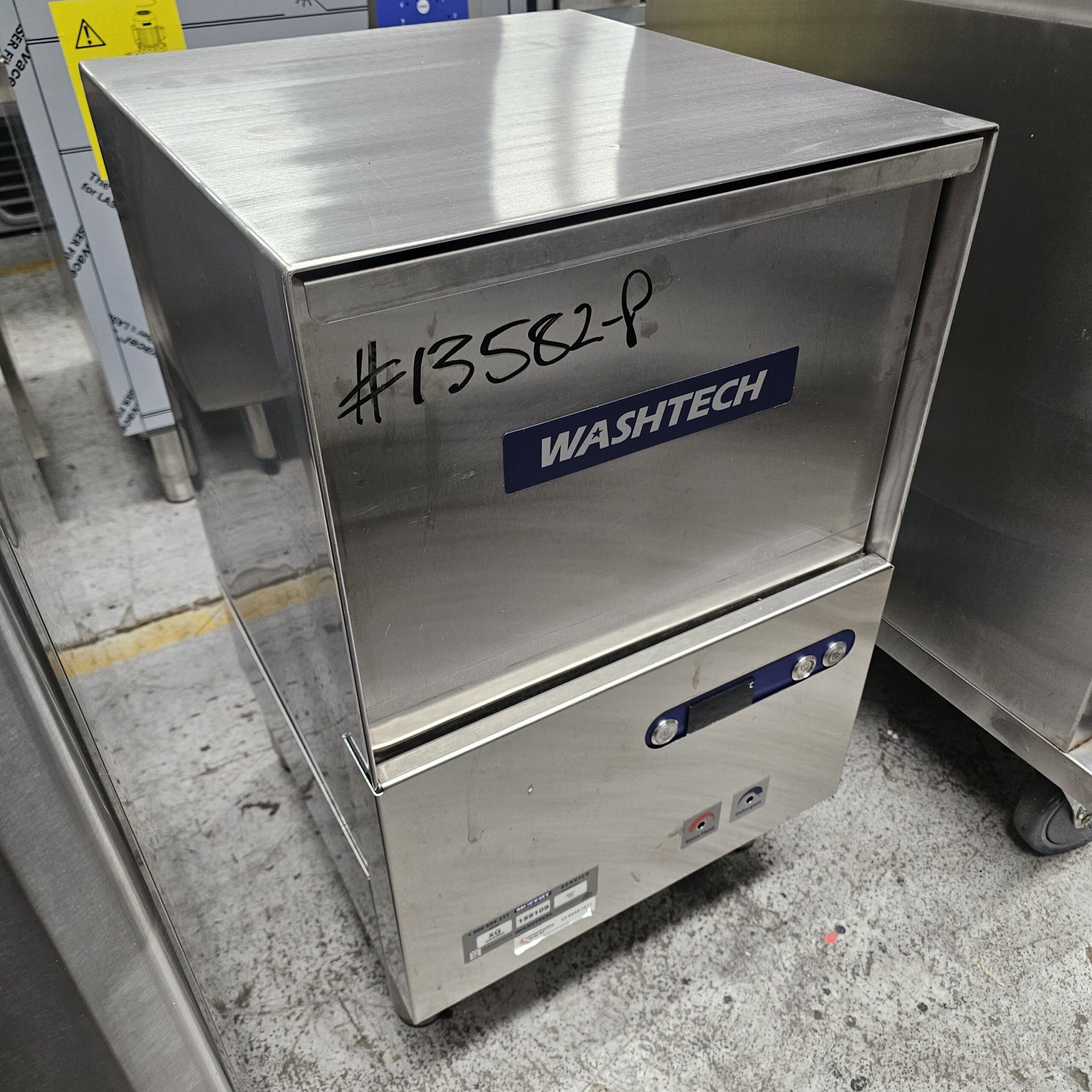 A stainless steel Washtech commercial dishwasher with a digital display and buttons sits on a concrete floor. #13582P is written in black marker on the top front panel. Other metal equipment is visible nearby. MRCE