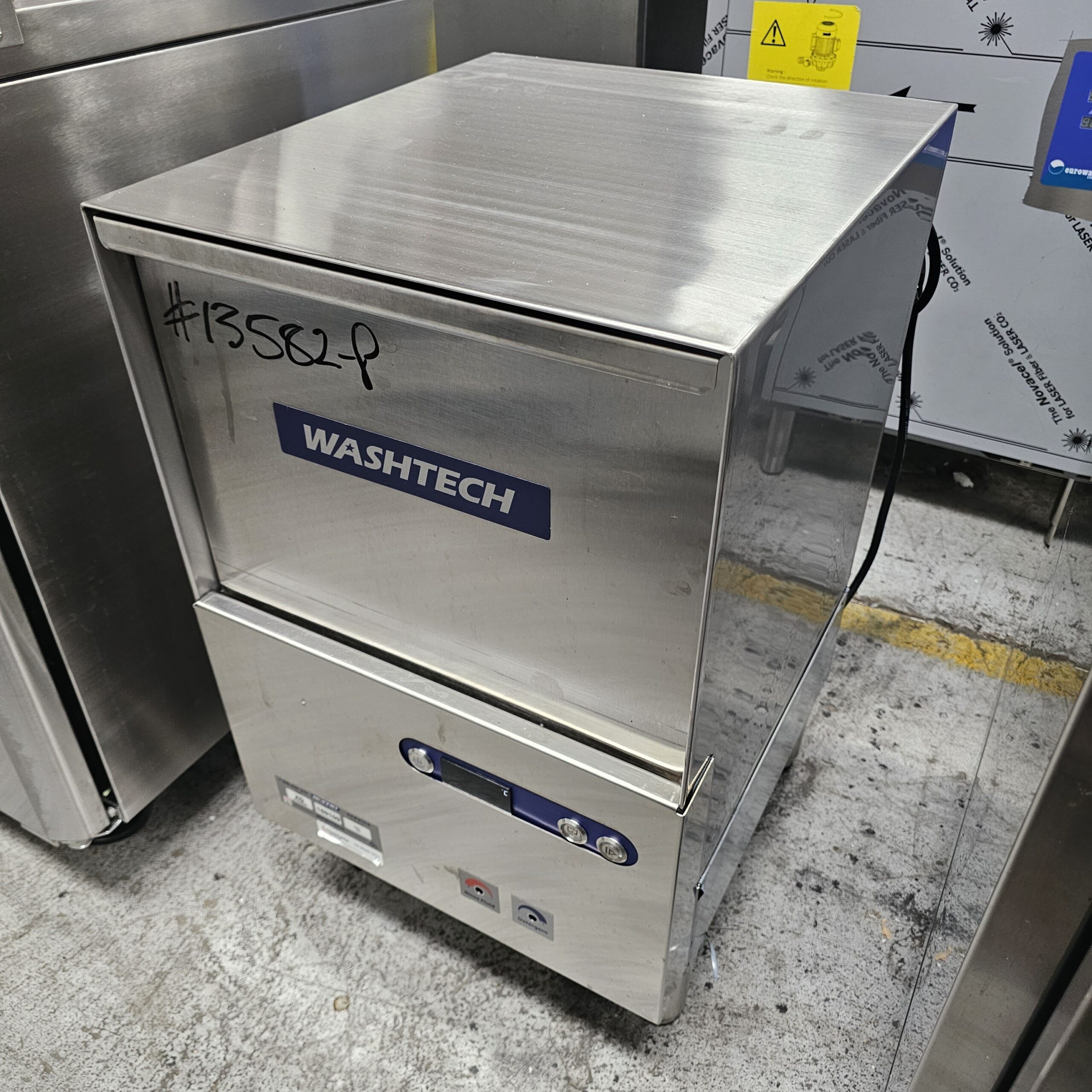 A stainless steel WASHTECH commercial dishwasher sits on a concrete floor, with the code #13582F written in black marker on the top left corner. Control buttons and indicator lights are visible on the front panel. MRCE