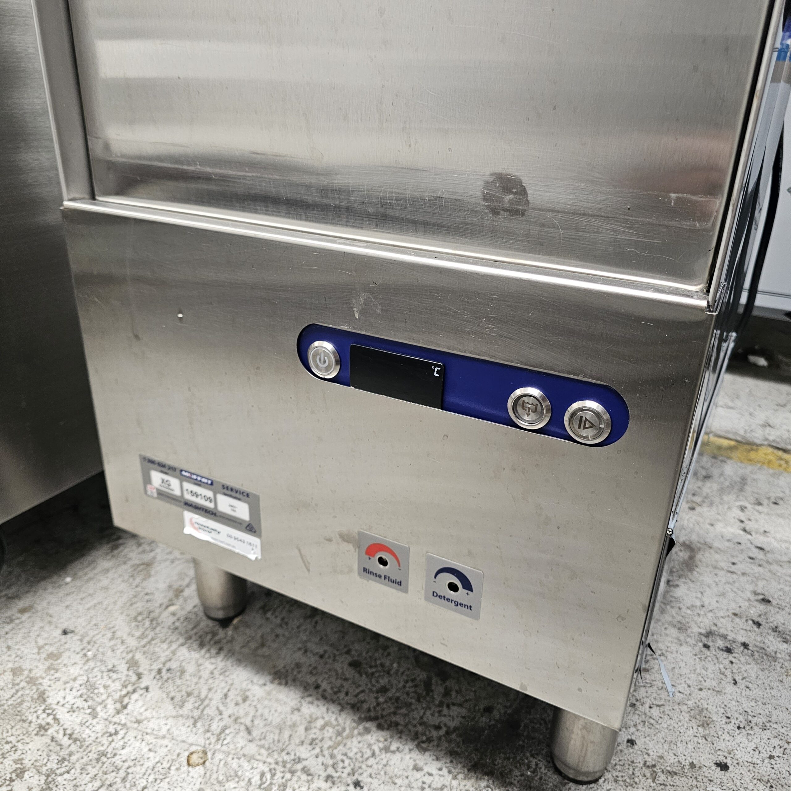 A stainless steel industrial machine with a blue control panel featuring a small digital display and two buttons. There are labels and stickers below the panel, and the unit stands on short metal legs on a concrete floor. MRCE