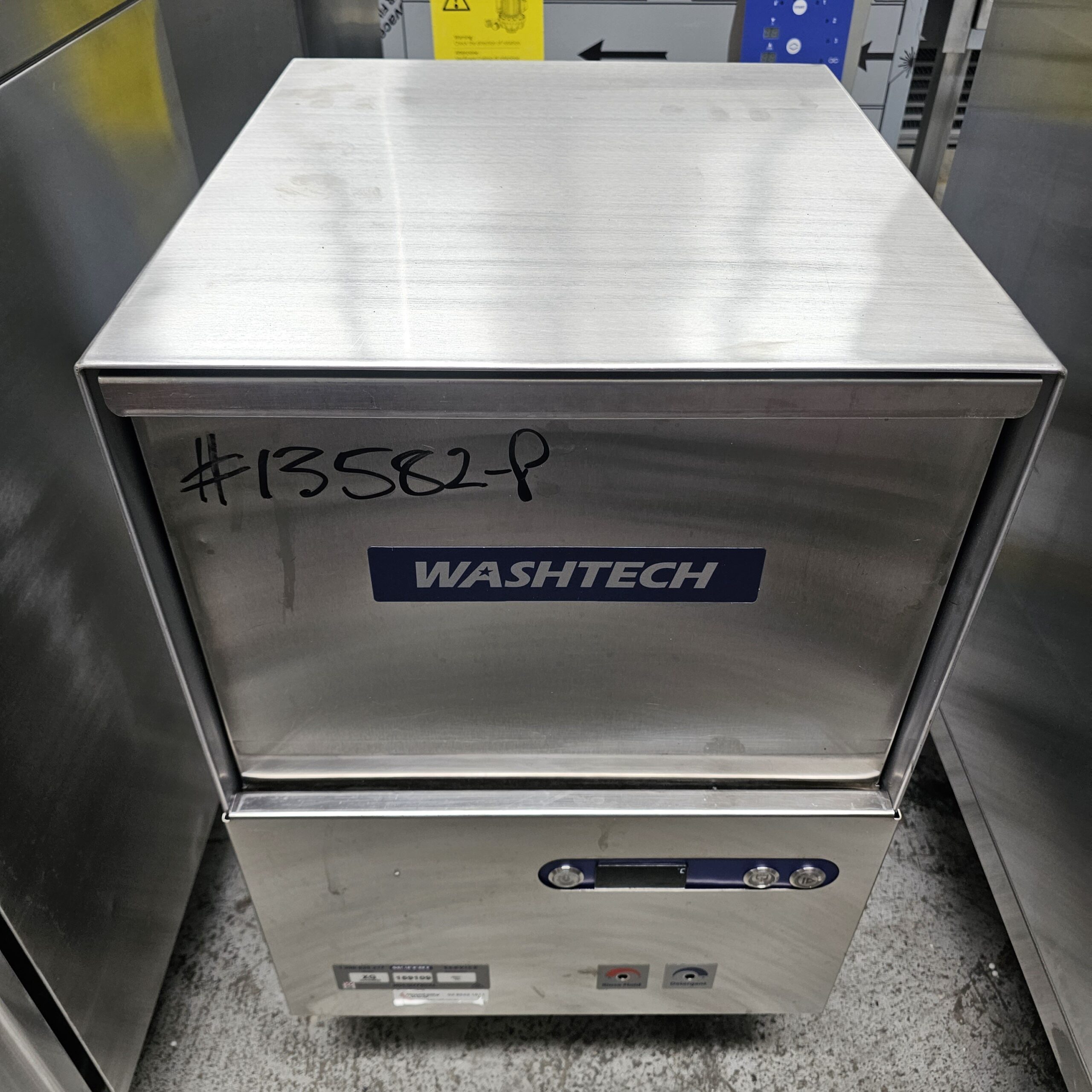 A stainless steel Washtech commercial dishwasher with the code #13582P written on the front, standing on a concrete floor in an industrial kitchen or storage area. MRCE