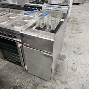 A commercial deep fryer with two wire baskets and blue handles sits next to a stainless steel oven in a kitchen appliance showroom. The fryer has a label and a handwritten number “13863P” on it. MRCE