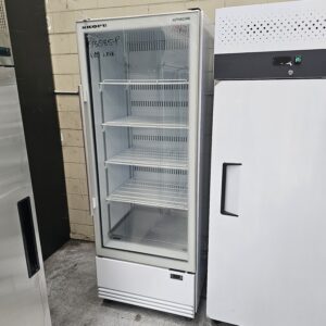 A glass-front commercial refrigerator with four wire shelves stands next to another white appliance in an industrial setting. SKOPE and 600 LITRE are written on the top left of the glass door. MRCE