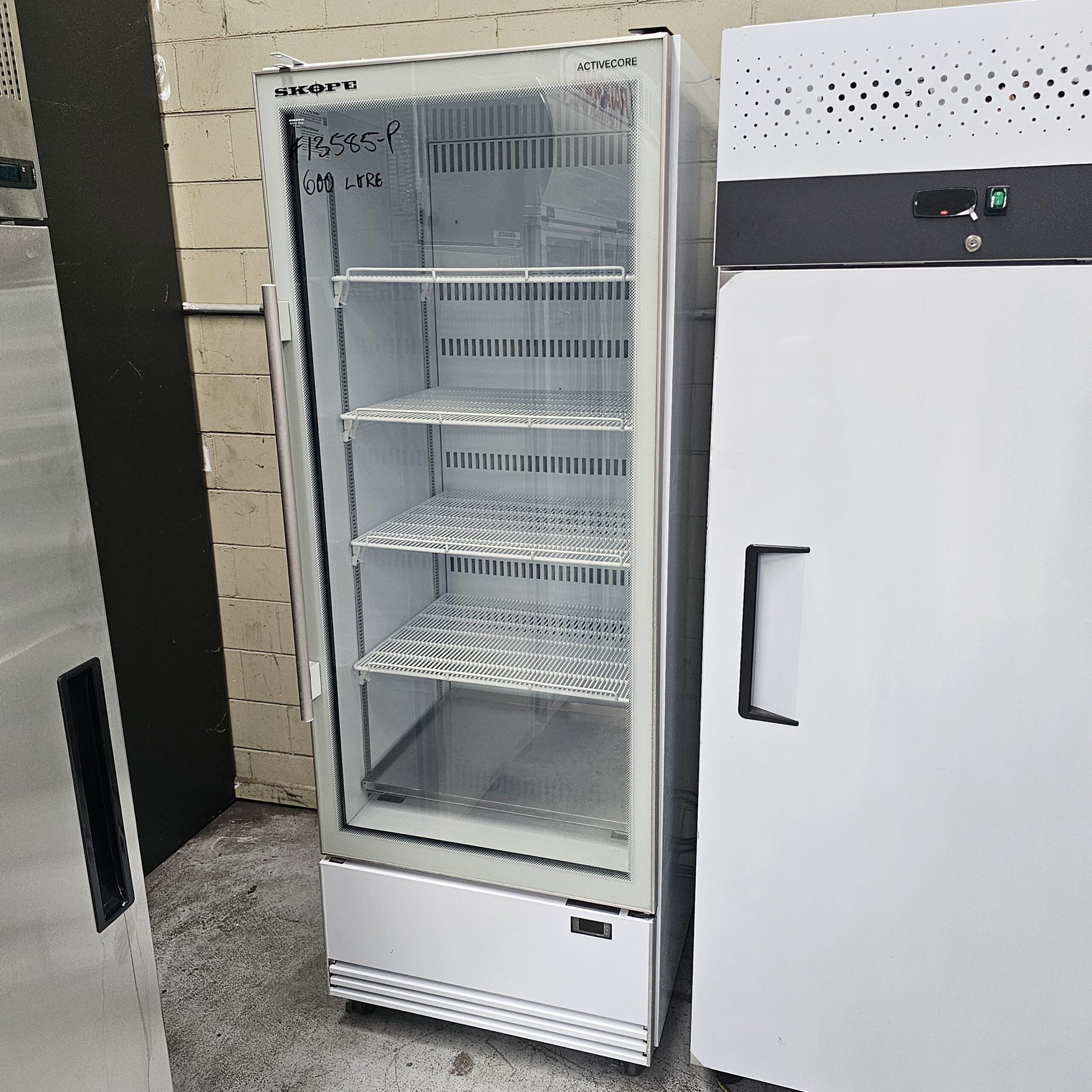 A glass-front commercial refrigerator with four wire shelves stands next to another white appliance in an industrial setting. SKOPE and 600 LITRE are written on the top left of the glass door. MRCE