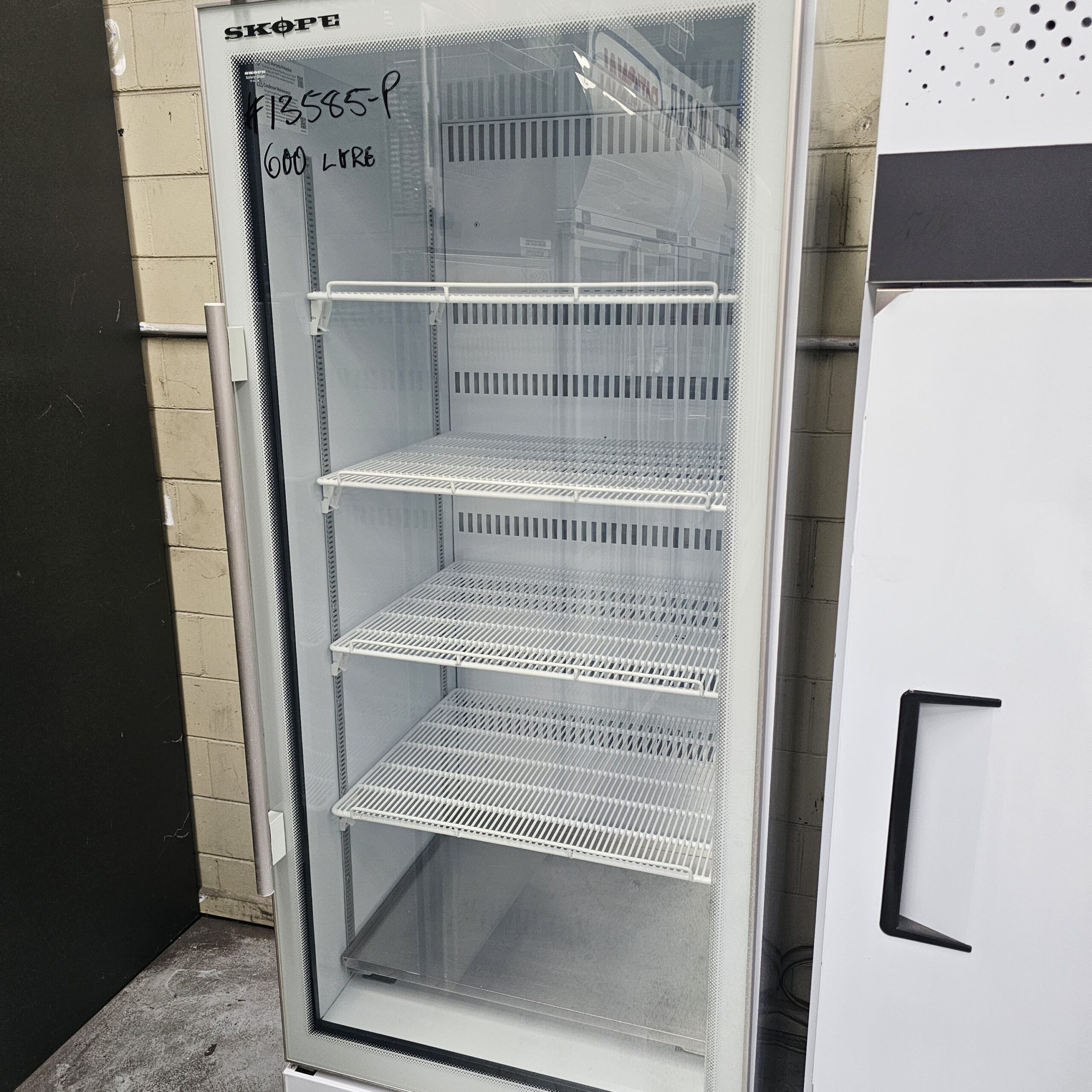 A tall, glass-fronted display fridge with four empty wire shelves stands against a tiled wall. “$2585-P 600 Litre” is handwritten on the top left of the glass door. Another appliance is partially visible to the right. MRCE