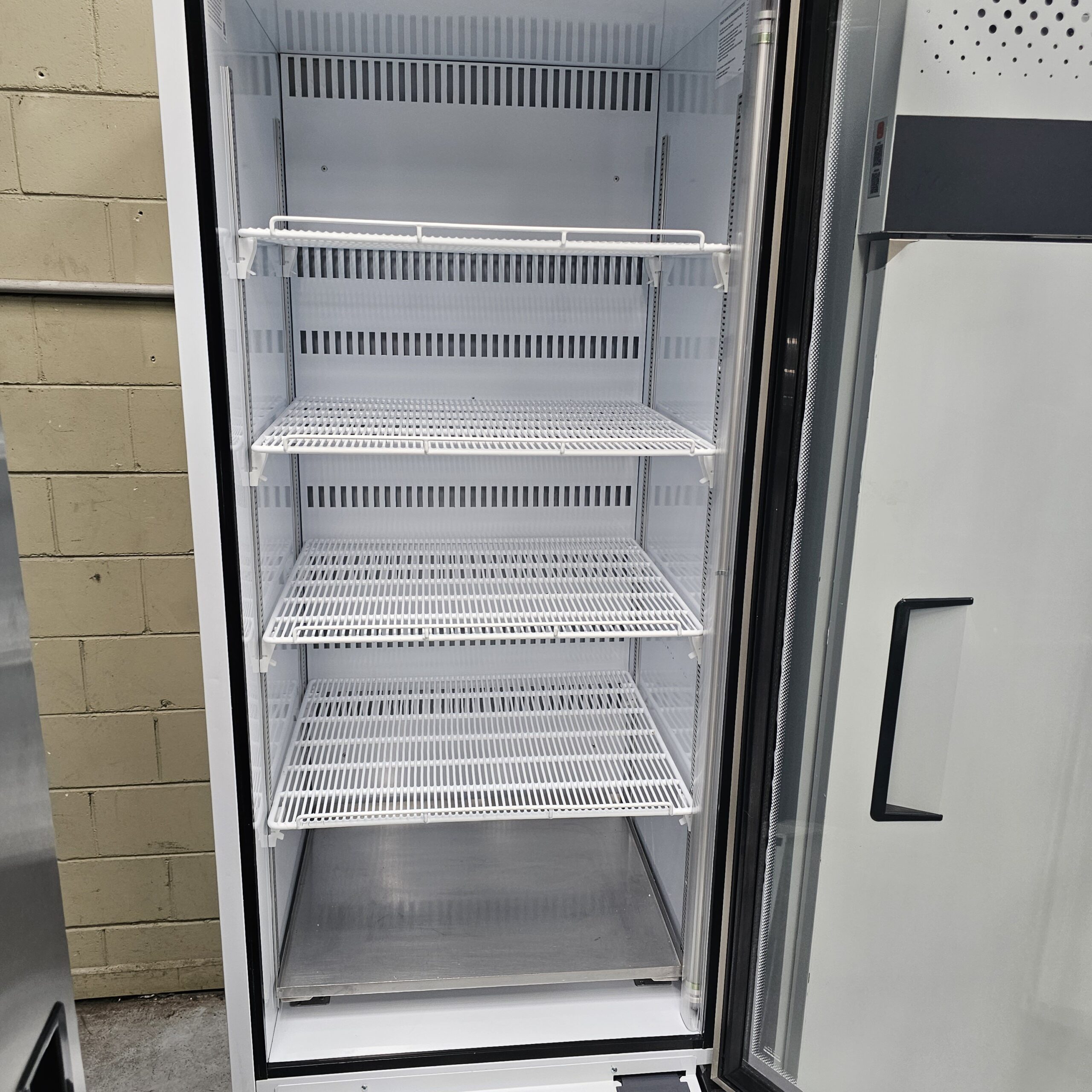 An empty commercial refrigerator with three white wire shelves, a glass door, and a beige brick wall visible on the left side. The refrigerator is clean and completely open. MRCE