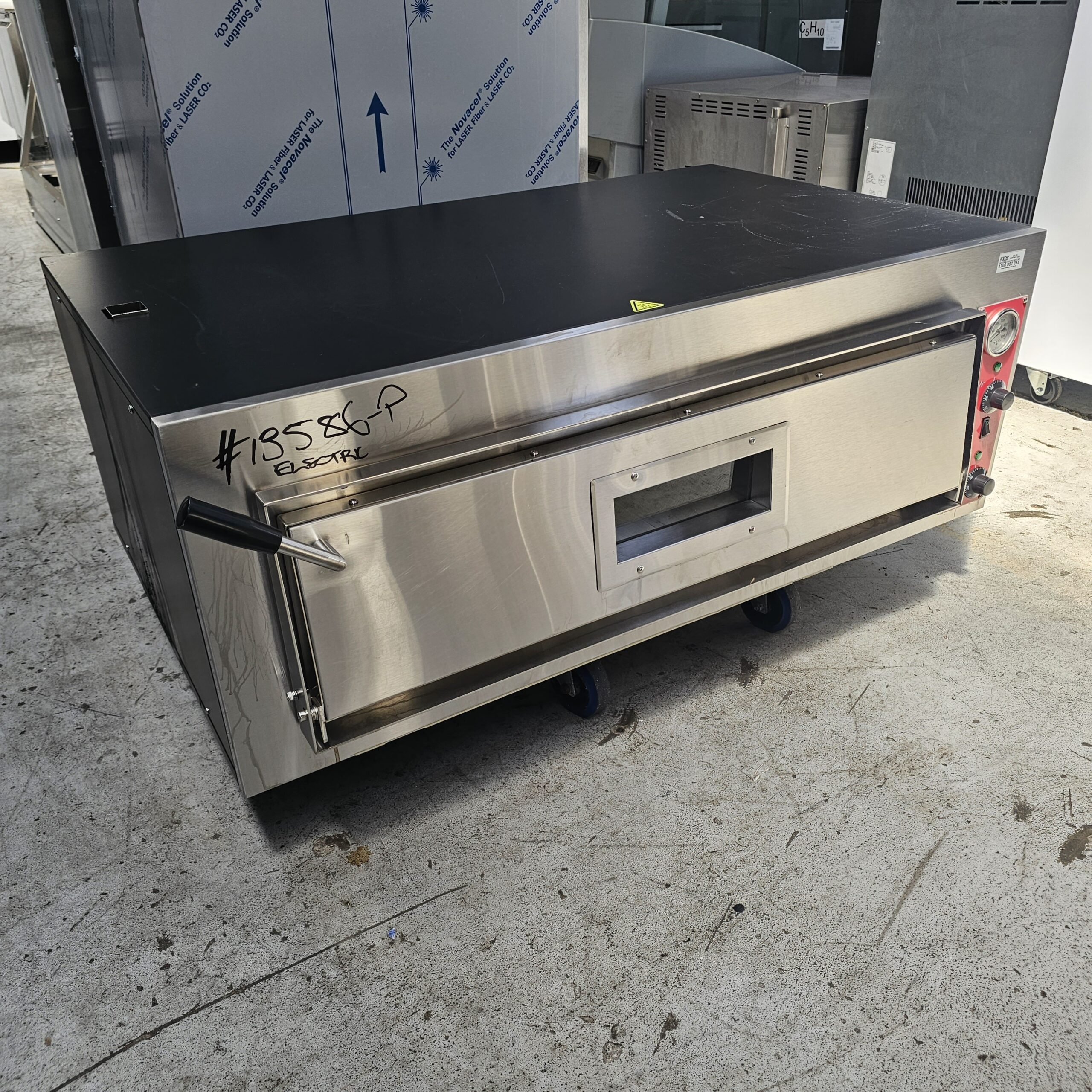A large stainless steel commercial oven sits on a concrete floor, with a rectangular front window and control knobs on the right side. A handwritten label reads #13580P Electric. MRCE