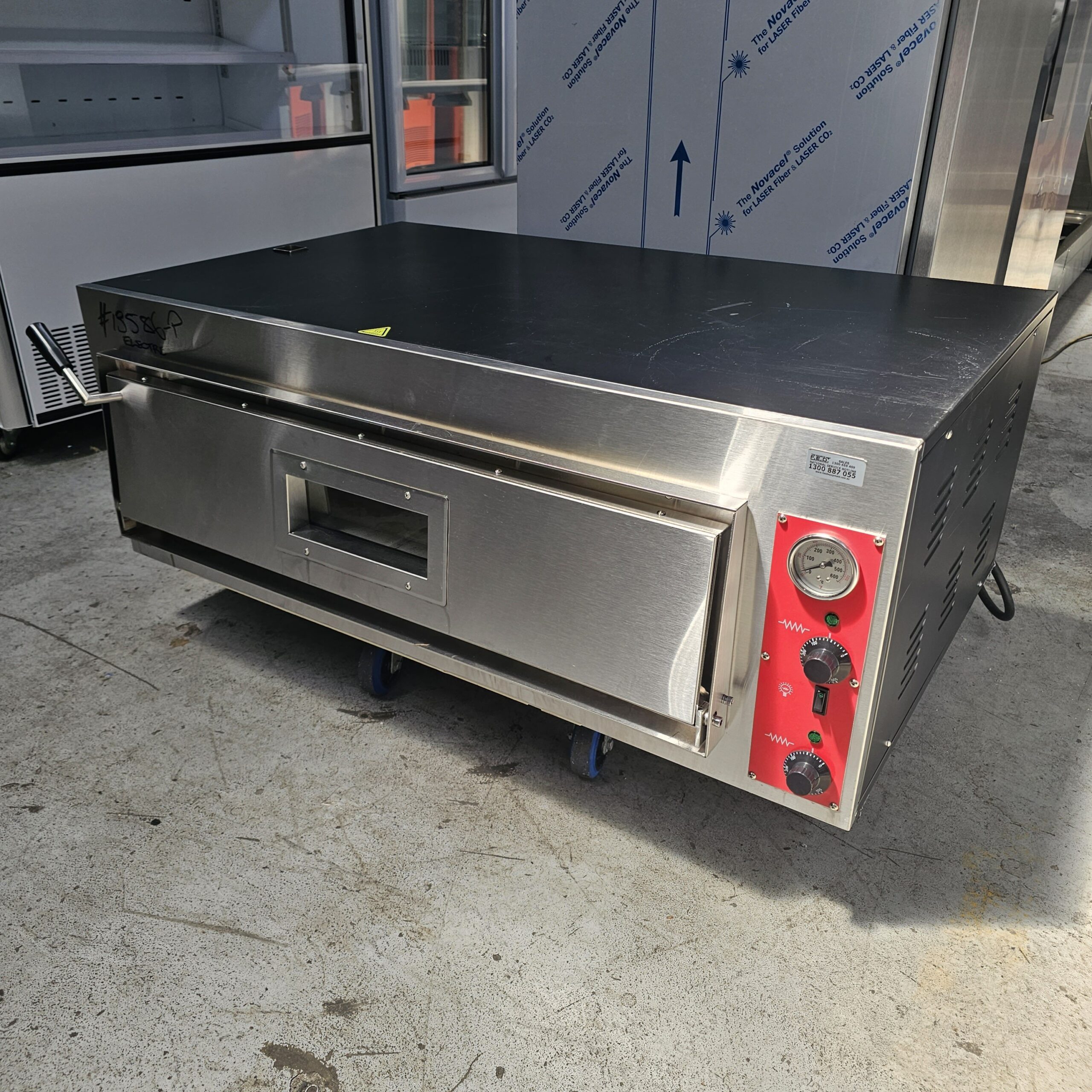 A stainless steel commercial pizza oven with a front window, control knobs, and a temperature gauge on the right side, sitting on a concrete floor in a commercial kitchen or appliance showroom. MRCE