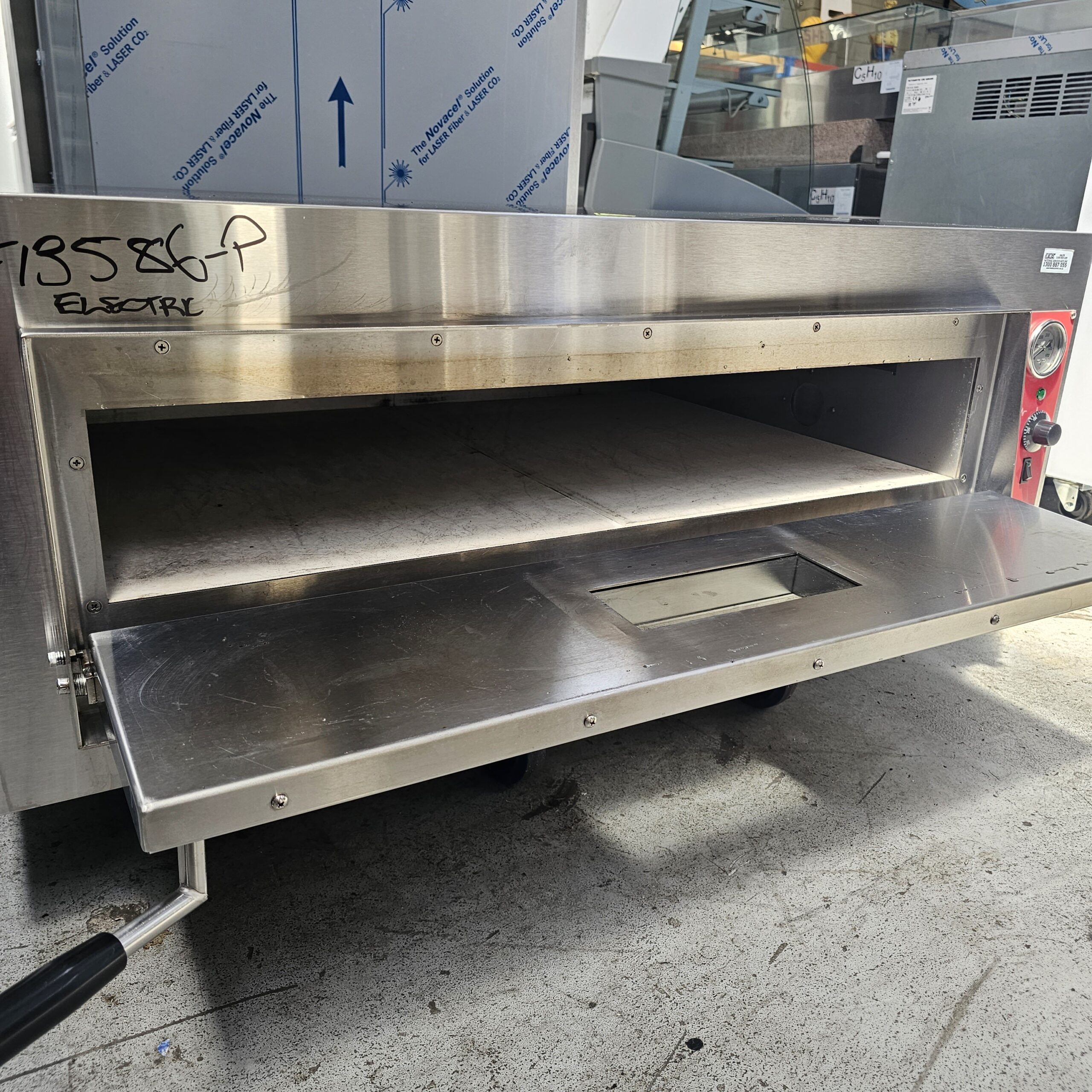 A commercial stainless steel pizza oven with an open front and a pull-down door, placed on a concrete floor in an industrial kitchen setting. MRCE