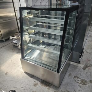 A commercial glass display case with multiple empty shelves is shown on a concrete floor, reflecting light from above. The display case has a stainless steel base and is situated in an industrial setting. MRCE