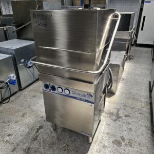 A commercial stainless steel Sharpline dishwasher with control buttons and dials on the front is placed in an industrial setting. The upper door is partially elevated, and handwritten notes are visible on the top. MRCE