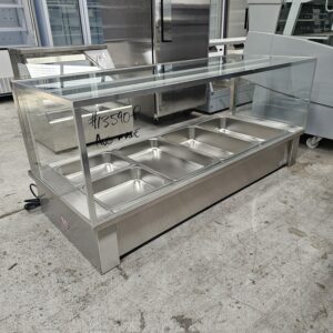 A stainless steel and glass food display counter with six empty metal food trays and a glass sneeze guard, situated in an industrial indoor setting with other equipment in the background. MRCE