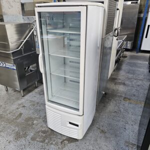A white, upright commercial display refrigerator with a glass door and empty shelves stands on a concrete floor in an industrial or warehouse setting among other metal kitchen equipment. MRCE