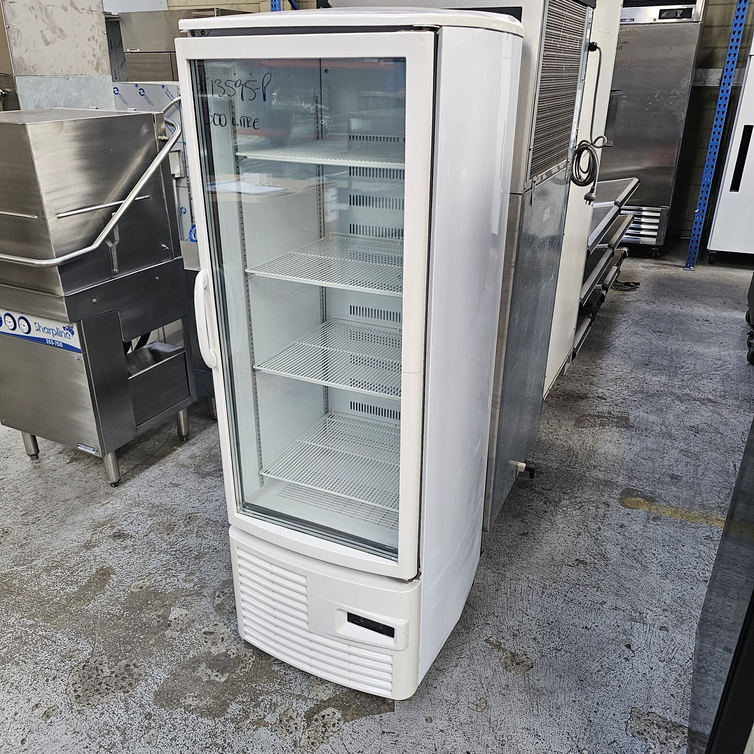 A white, upright commercial display refrigerator with a glass door and empty shelves stands on a concrete floor in an industrial or warehouse setting among other metal kitchen equipment. MRCE