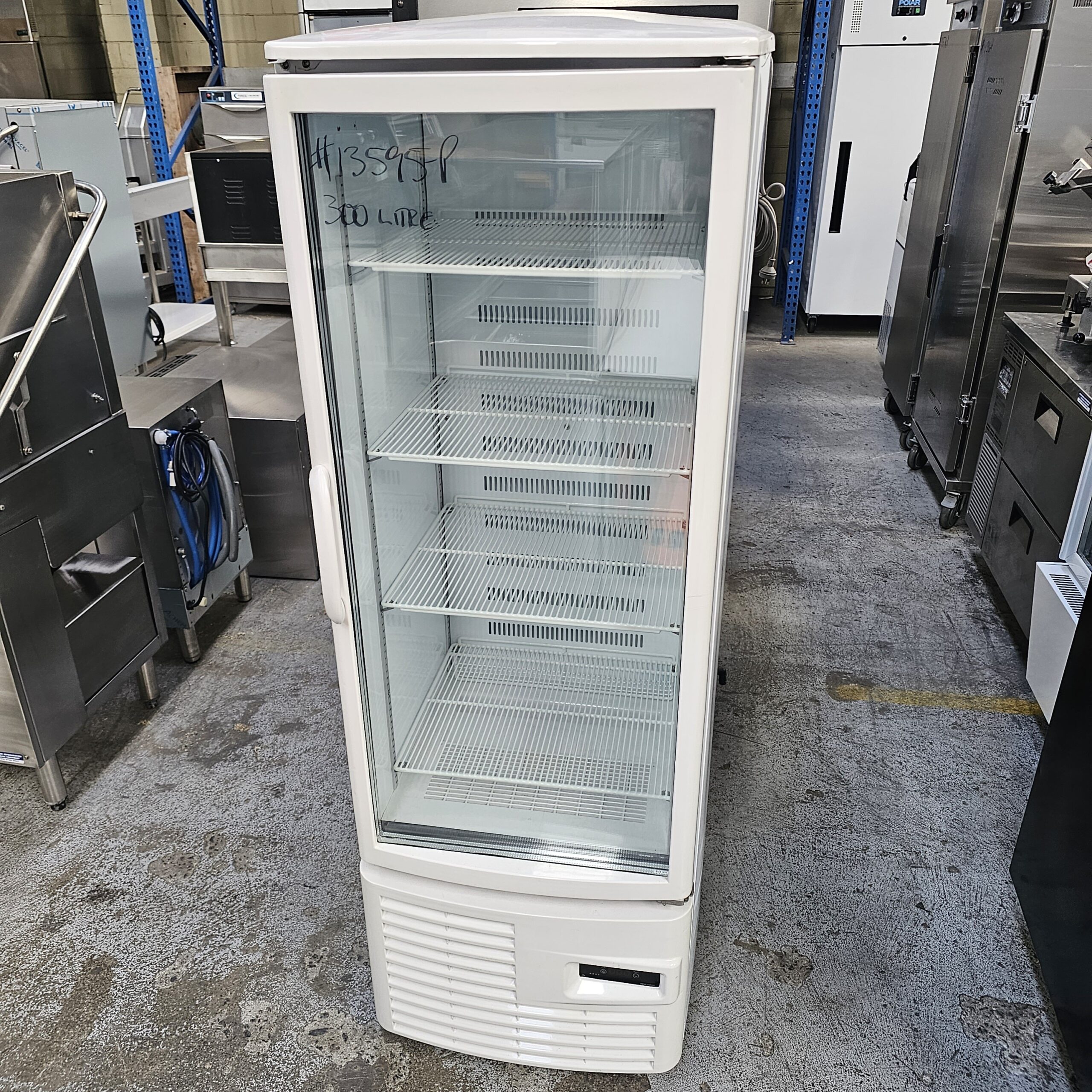 A tall, white commercial display refrigerator with a glass door and multiple empty shelves stands in an industrial setting with other kitchen equipment around it. Handwritten text is visible on the glass door. MRCE