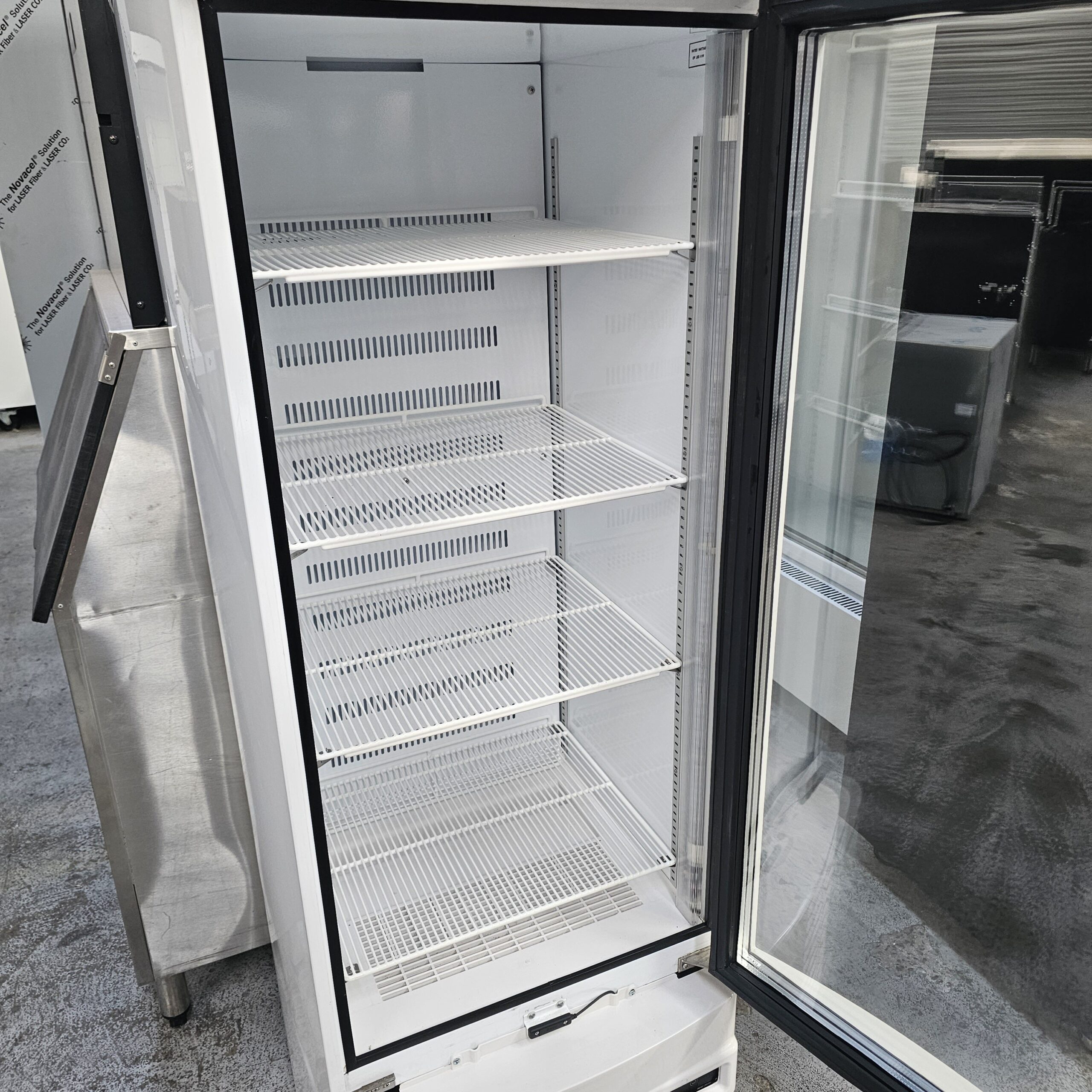 An empty commercial refrigerator with a glass door is open, displaying several white wire shelves inside. The fridge is situated in an industrial or warehouse setting. MRCE