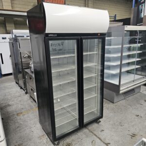 A commercial double-door glass display refrigerator labeled APOLLO stands on a warehouse floor, with empty shelves inside and other appliances visible in the background. MRCE
