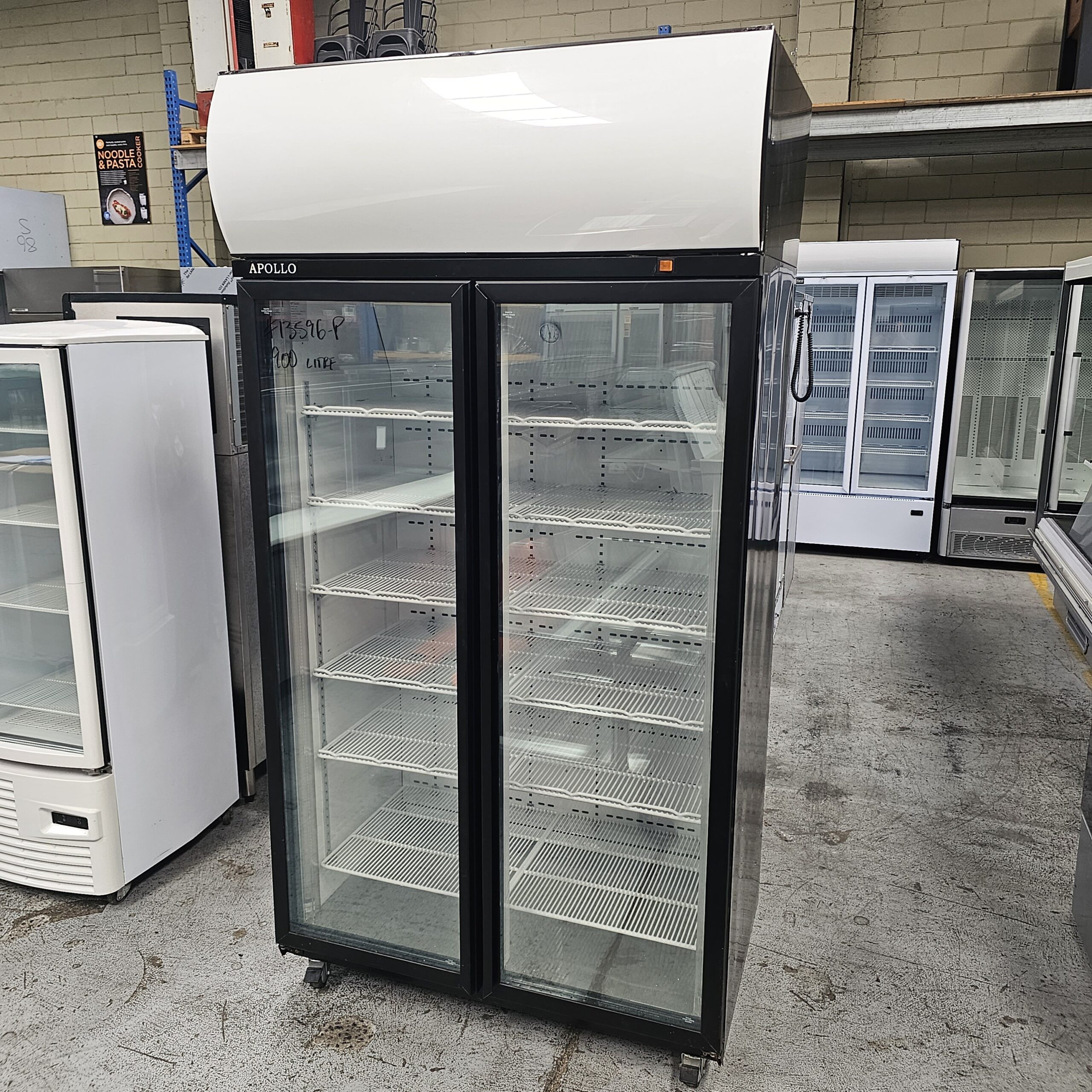 A commercial glass-door refrigerator with two sections and empty shelves stands in a warehouse with other refrigerators and freezers in the background. MRCE