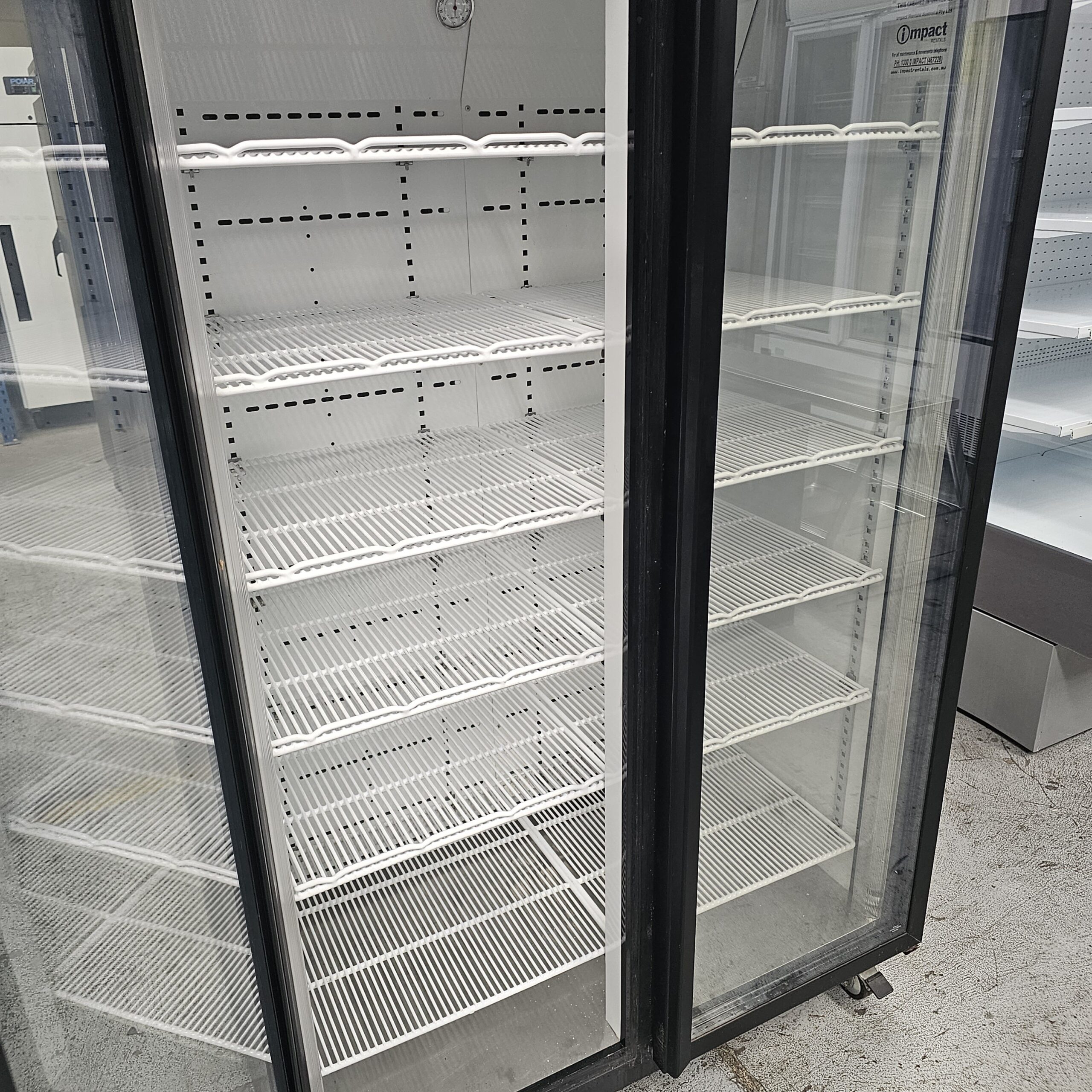 An open commercial refrigerator with glass doors and several empty white wire shelves inside, located in a store or warehouse setting. MRCE