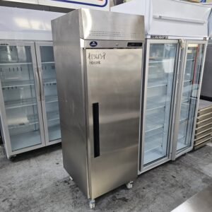 A tall, stainless steel commercial freezer with a black handle stands on wheels in front of other glass-front refrigerators inside a store or warehouse. A label reads #13639 FREEZER. MRCE