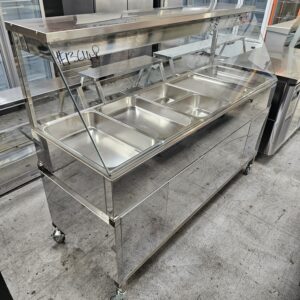 A stainless steel commercial food service counter with a glass sneeze guard, featuring multiple empty food trays, on wheels, placed on a concrete floor. MRCE