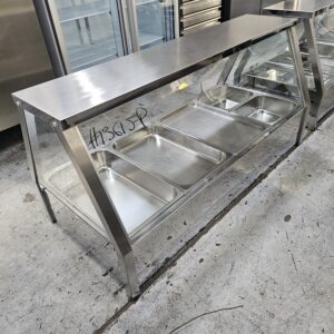 A stainless steel commercial food serving station with five empty metal trays underneath a glass sneeze guard, situated on a concrete floor in an industrial kitchen setting. MRCE