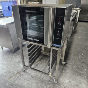 A commercial Turbofan convection oven on a stainless steel stand with multiple tray slots, situated in an industrial kitchen with metal countertops and equipment visible in the background. MRCE