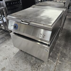 A large stainless steel commercial kitchen appliance, likely a tilting pan or bratt pan, sits on a concrete floor among other industrial kitchen equipment. The appliance has a blue dial and Electrolux branding. MRCE