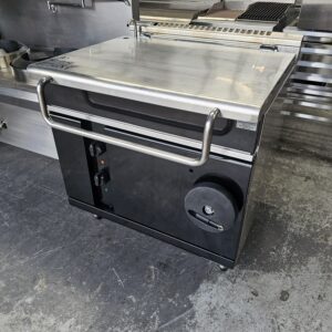 A commercial kitchen appliance with a closed stainless steel lid, black body, control panel, and a large round knob, standing on a concrete floor next to other metal kitchen equipment. MRCE