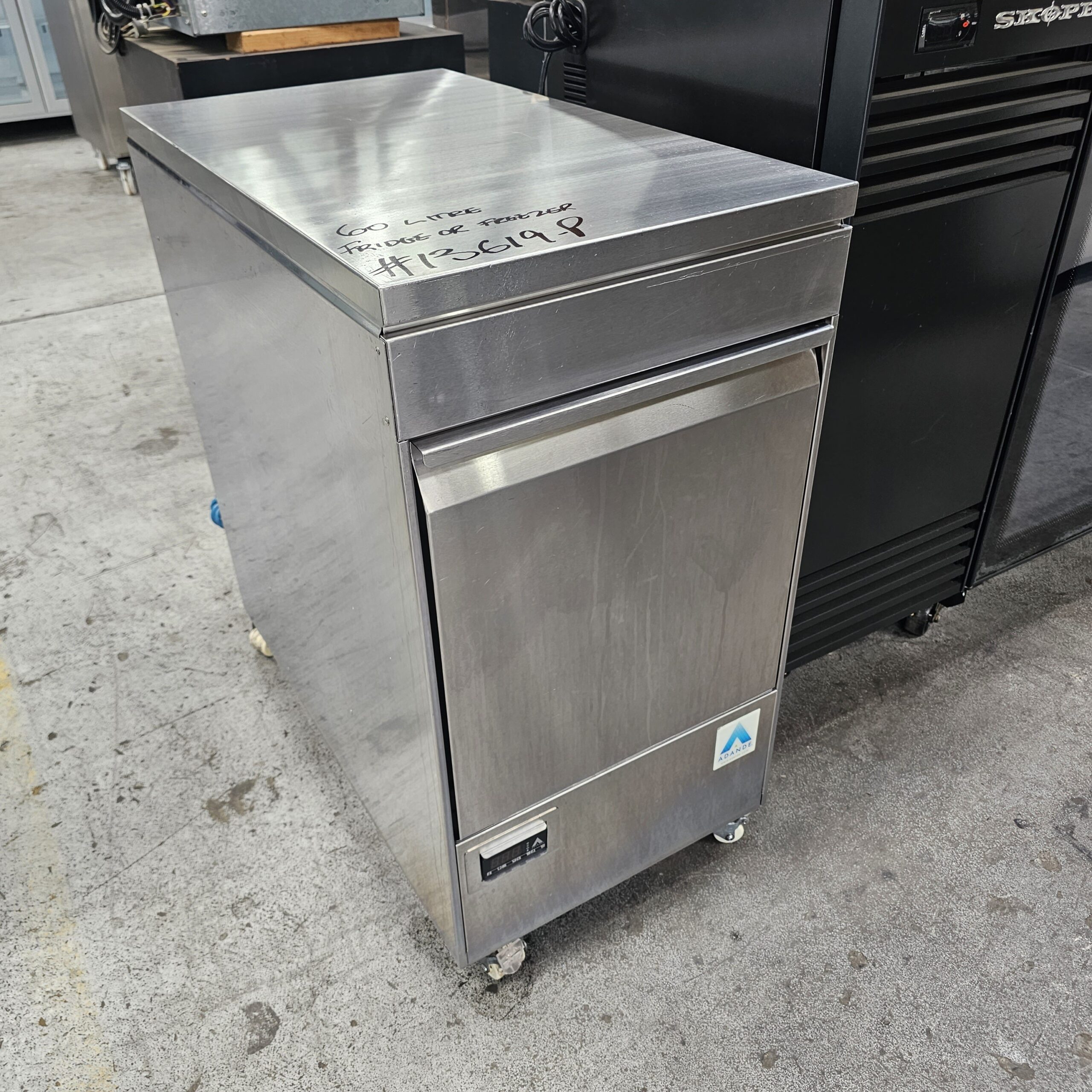 A stainless steel commercial refrigerator with a single door on wheels is positioned on a gray concrete floor, next to a black appliance. Handwritten text and a label are visible on its top surface. MRCE
