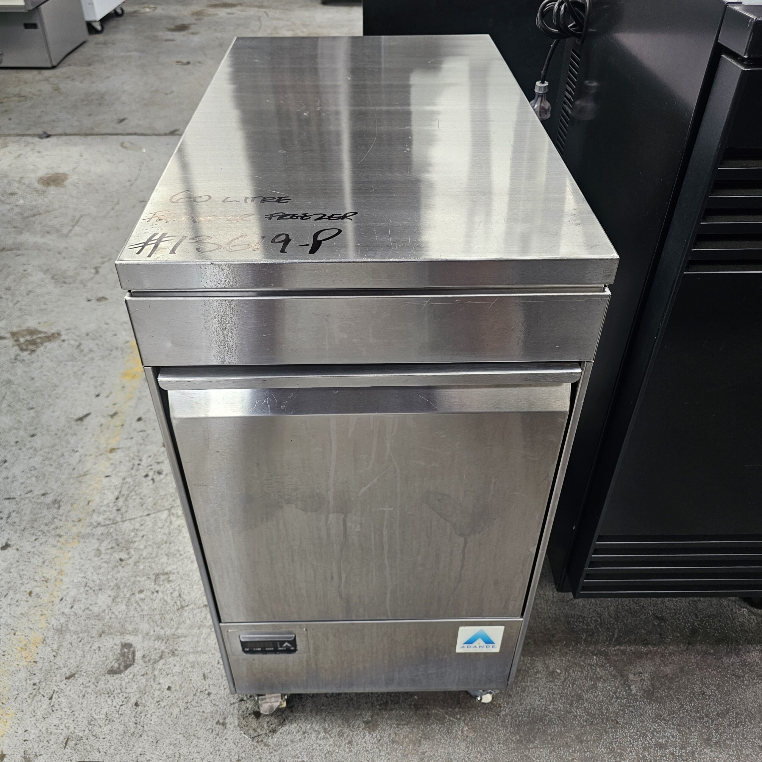 A stainless steel commercial refrigerator with a single door, set on four wheels, is placed on a concrete floor in an industrial setting. Handwritten markings and a label are visible on its top and front. MRCE