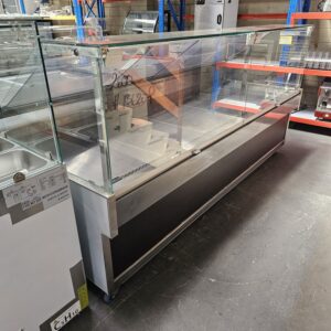 A large glass display case with metal frame sits on a concrete floor in a warehouse setting, surrounded by shelves and various equipment. A handwritten price is visible on the front glass. MRCE