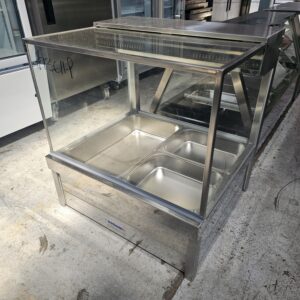 A stainless steel food display warmer with a glass top, containing four empty food trays, is standing on a concrete floor in a commercial kitchen setting. MRCE