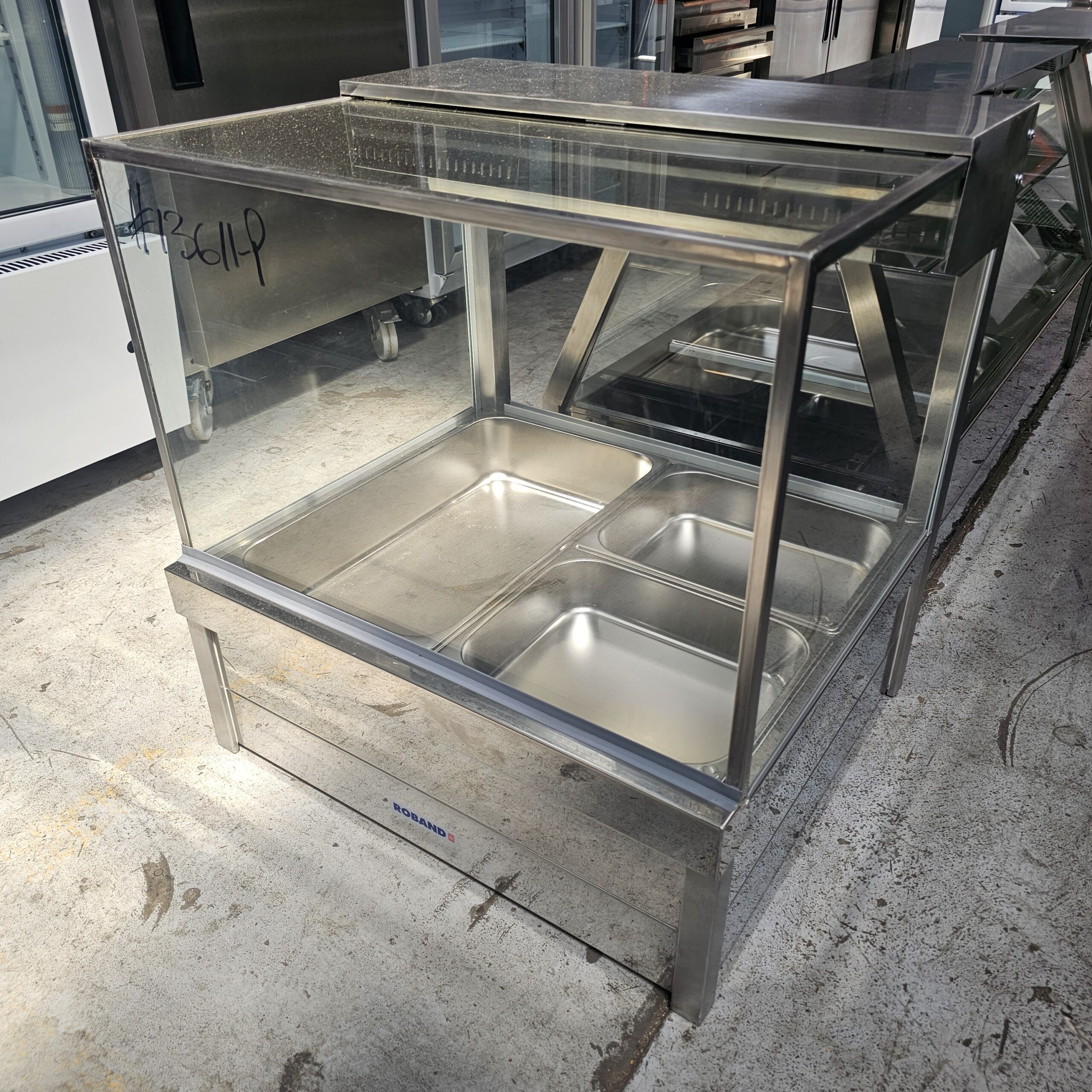 A stainless steel food display warmer with a glass top, containing four empty food trays, is standing on a concrete floor in a commercial kitchen setting. MRCE