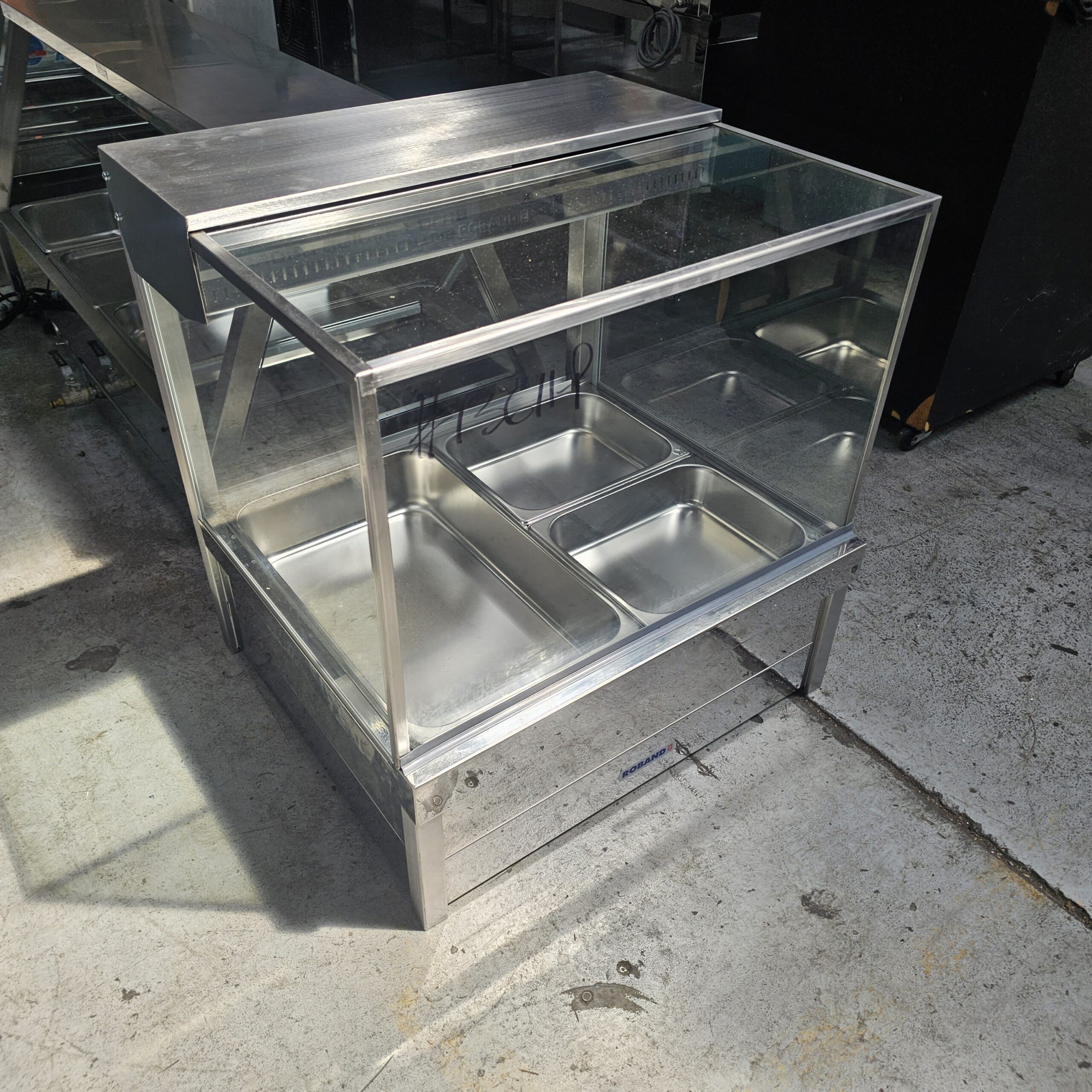 A stainless steel food display counter with a glass cover, featuring four empty metal trays inside, standing on a concrete floor in a well-lit indoor space. MRCE