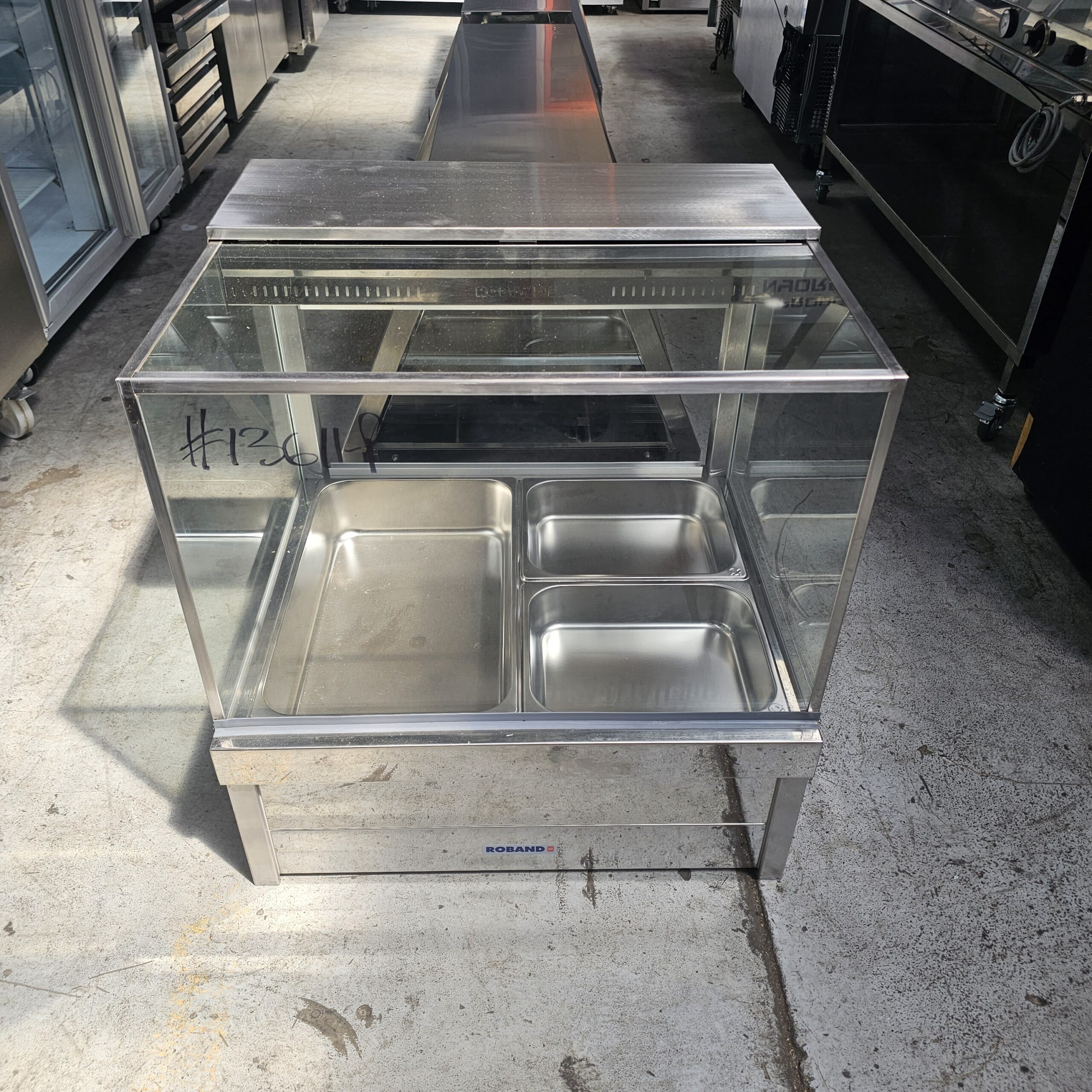 A stainless steel food display case with a glass front and top, containing three metal food trays, stands on a concrete floor in an industrial kitchen or equipment storage area. #126149 is written on the glass. MRCE