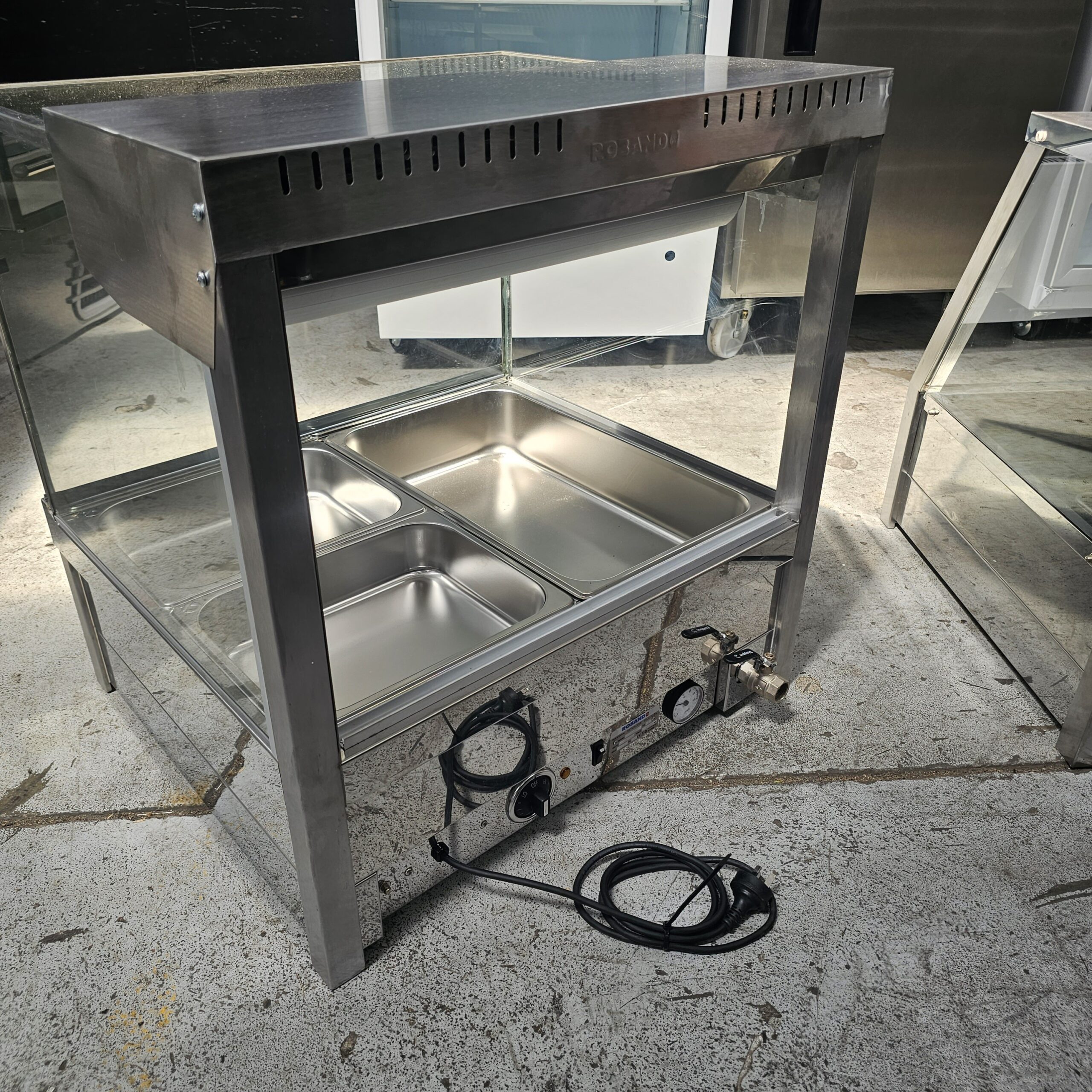 A stainless steel food warmer with two compartments on a lower shelf, temperature control knobs, a power cord, and a glass panel, standing on a concrete floor in a commercial kitchen setting. MRCE